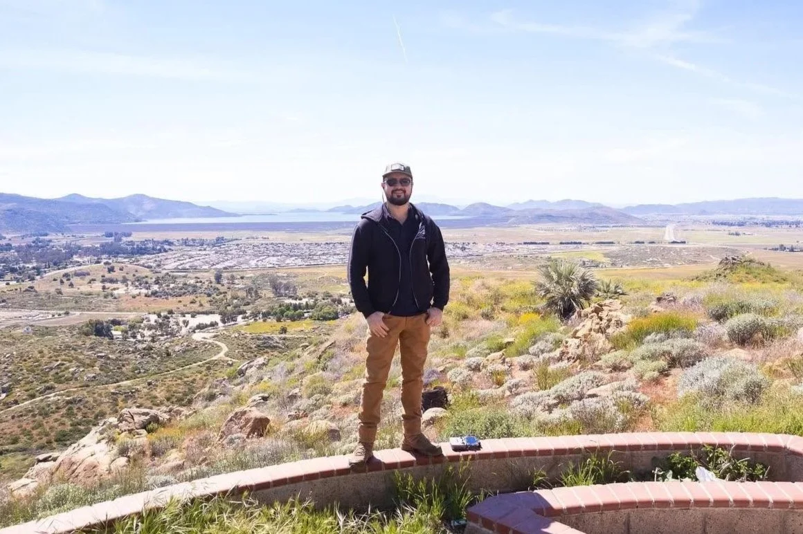 A man standing on a stone border overlooking a valley with hills, water, and distant mountains on a clear day.