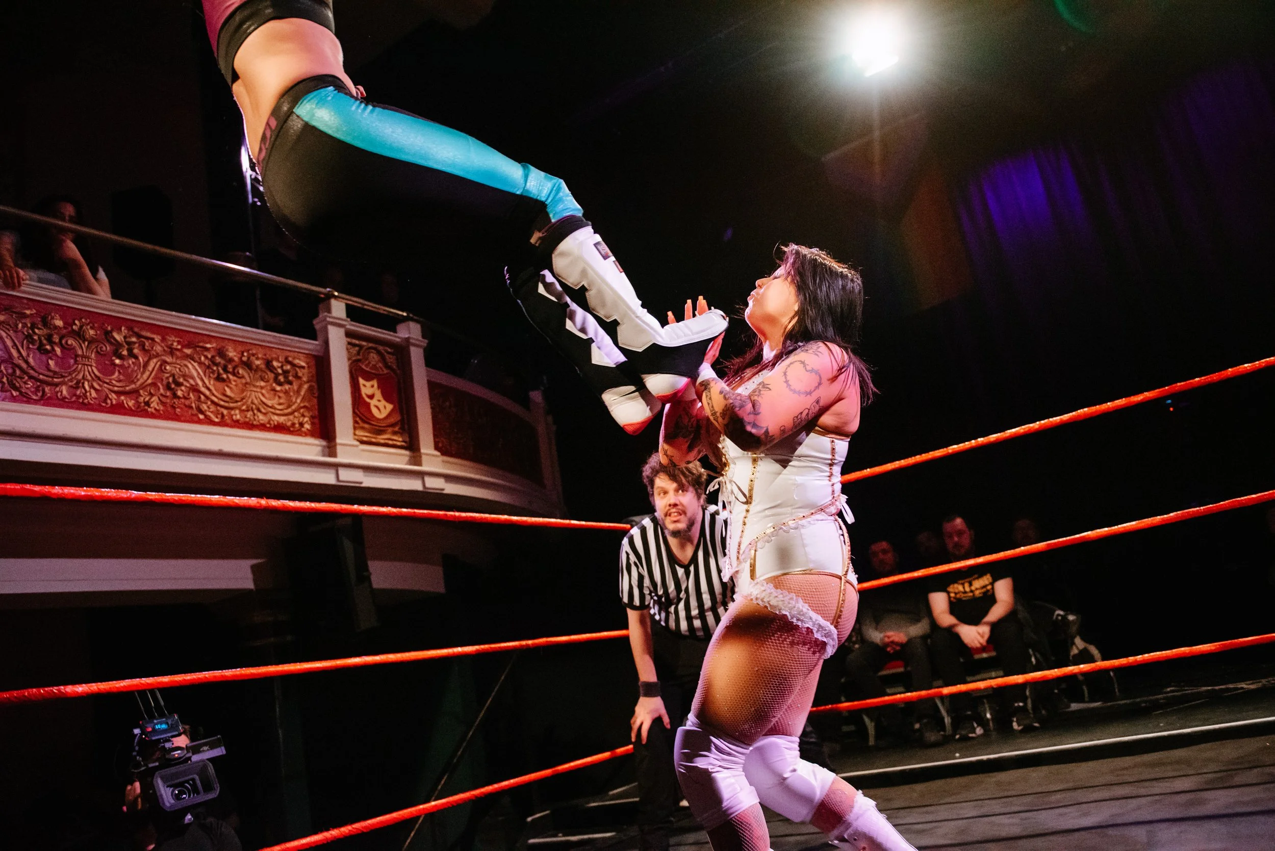 A professional female wrestler performing a move in a wrestling ring, with an audience and a referee watching.
