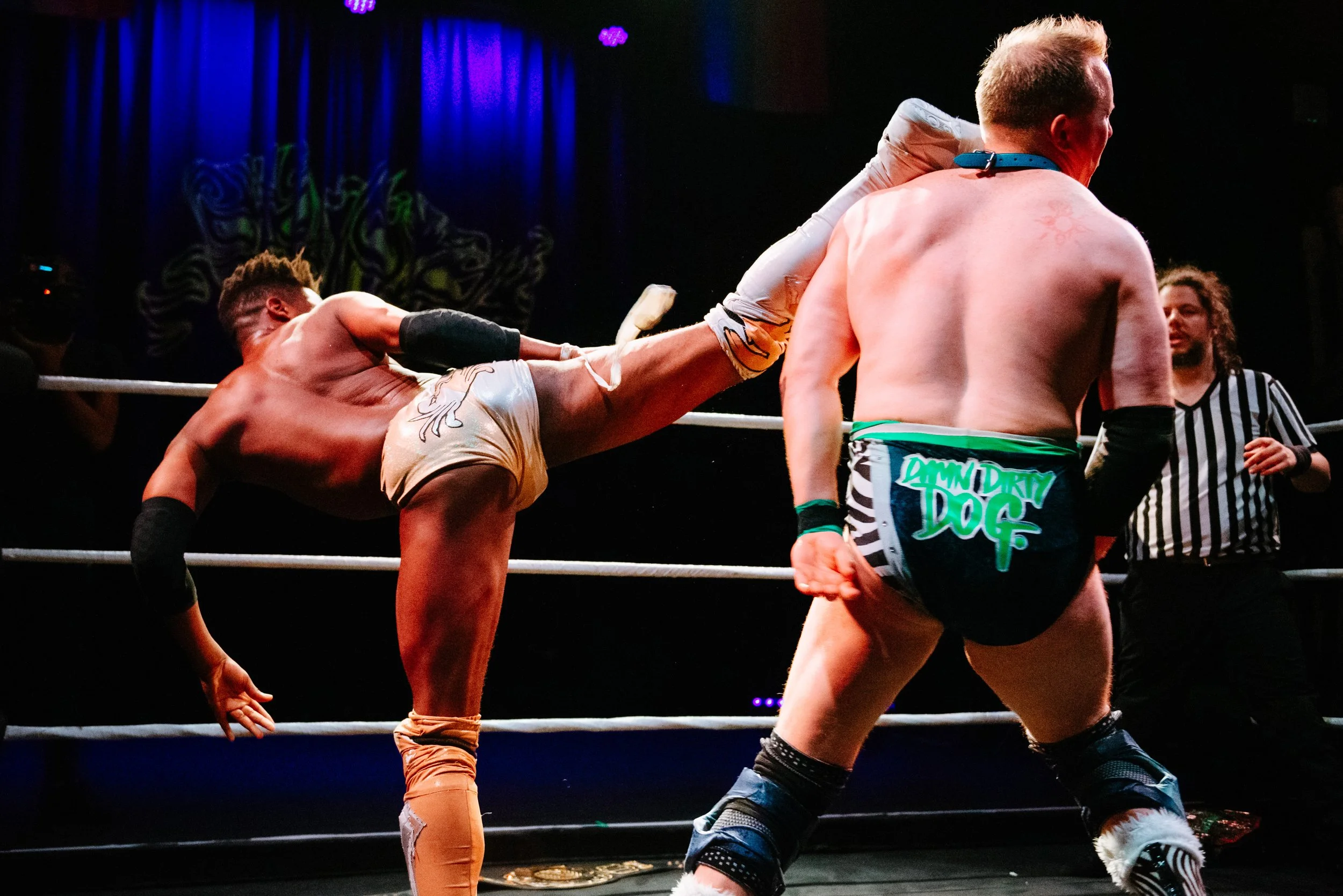 Two wrestlers in a wrestling ring, one performing a high kick on the other, with a referee observing in the background.