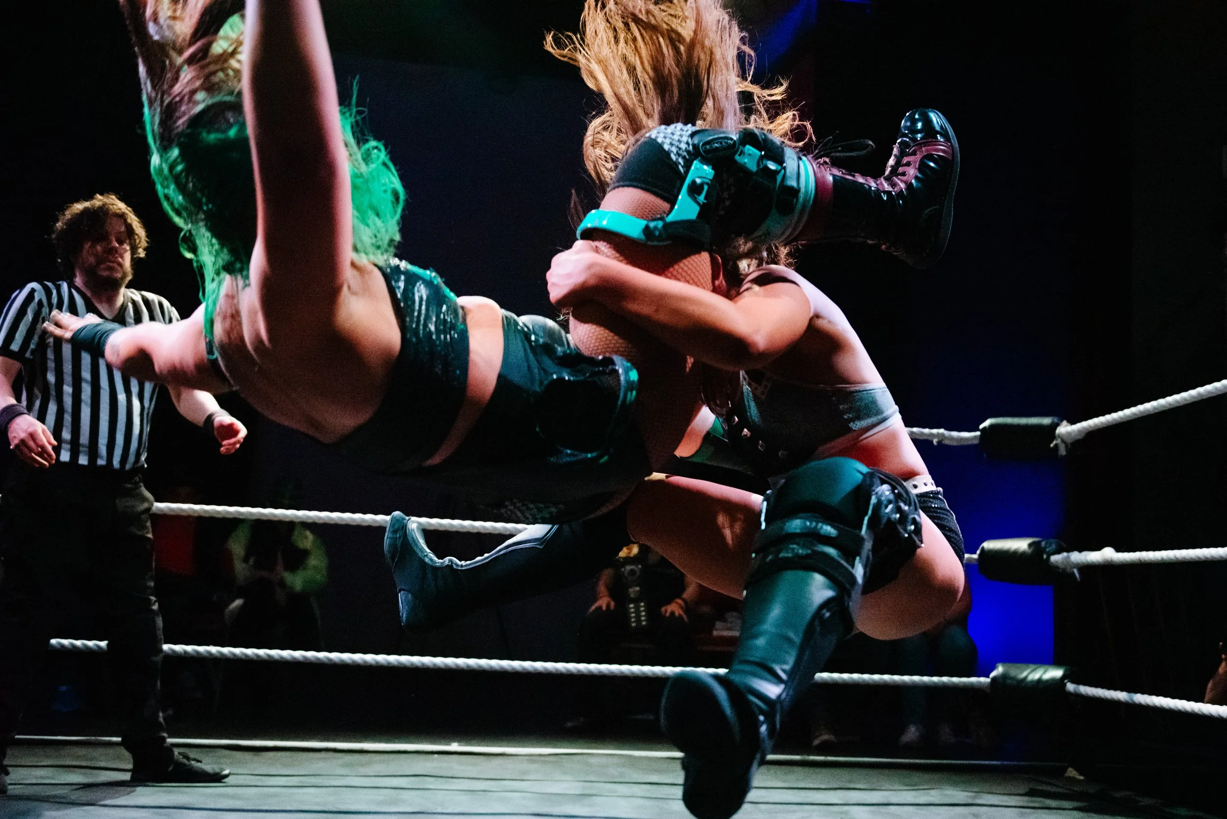 Two women wrestling in a ring with a referee watching, one woman has green hair and the other has long hair, is wearing knee-high boots.