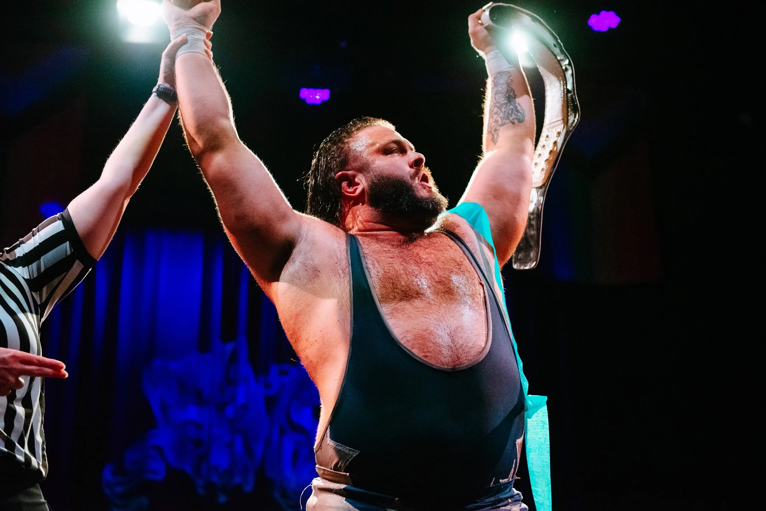 A victorious wrestler with a beard and tattoos, holding a championship belt above his head in a wrestling ring, with a referee touching his arm in celebration.