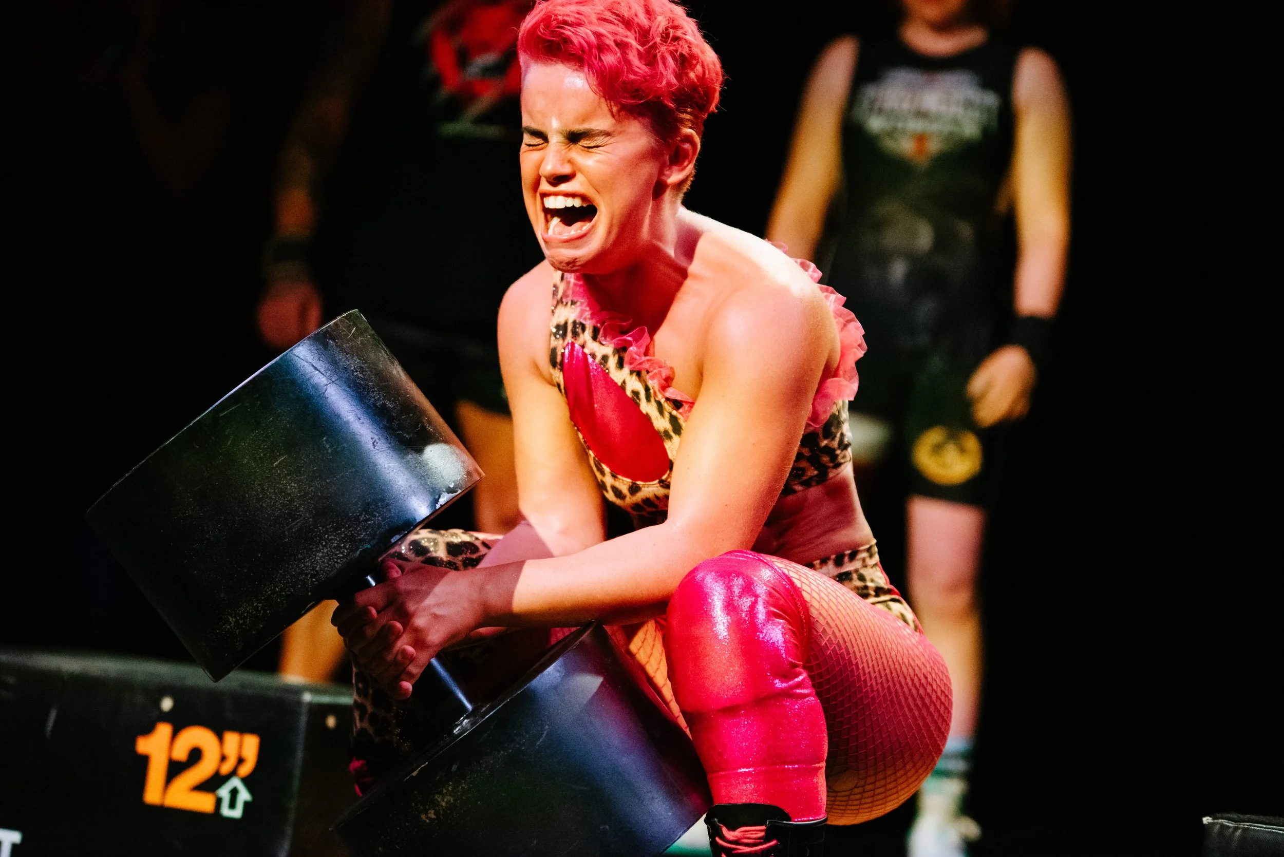 A woman with short red hair, wearing a leopard print and pink costume, is crouched down and appears to be in an emotional moment, holding a large black container. She is wearing pink fishnet tights and black shoes. The background shows others in simi