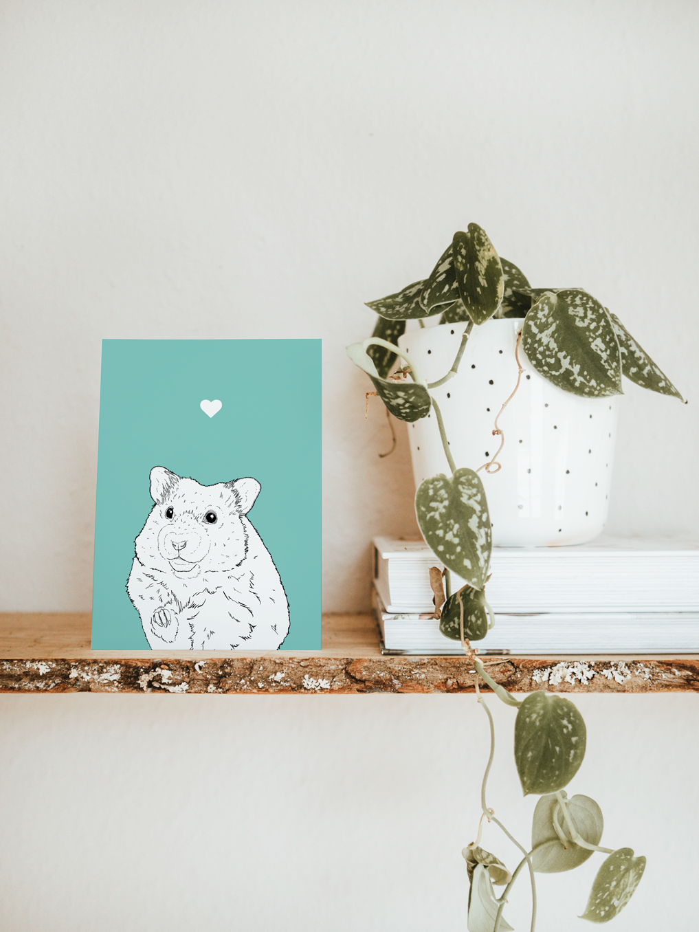 An A6 greetings card & white plant pot on a shelf. The card has a pale blue coloured background, with a black & white digital illustration of the head & shoulders of a Hamster.