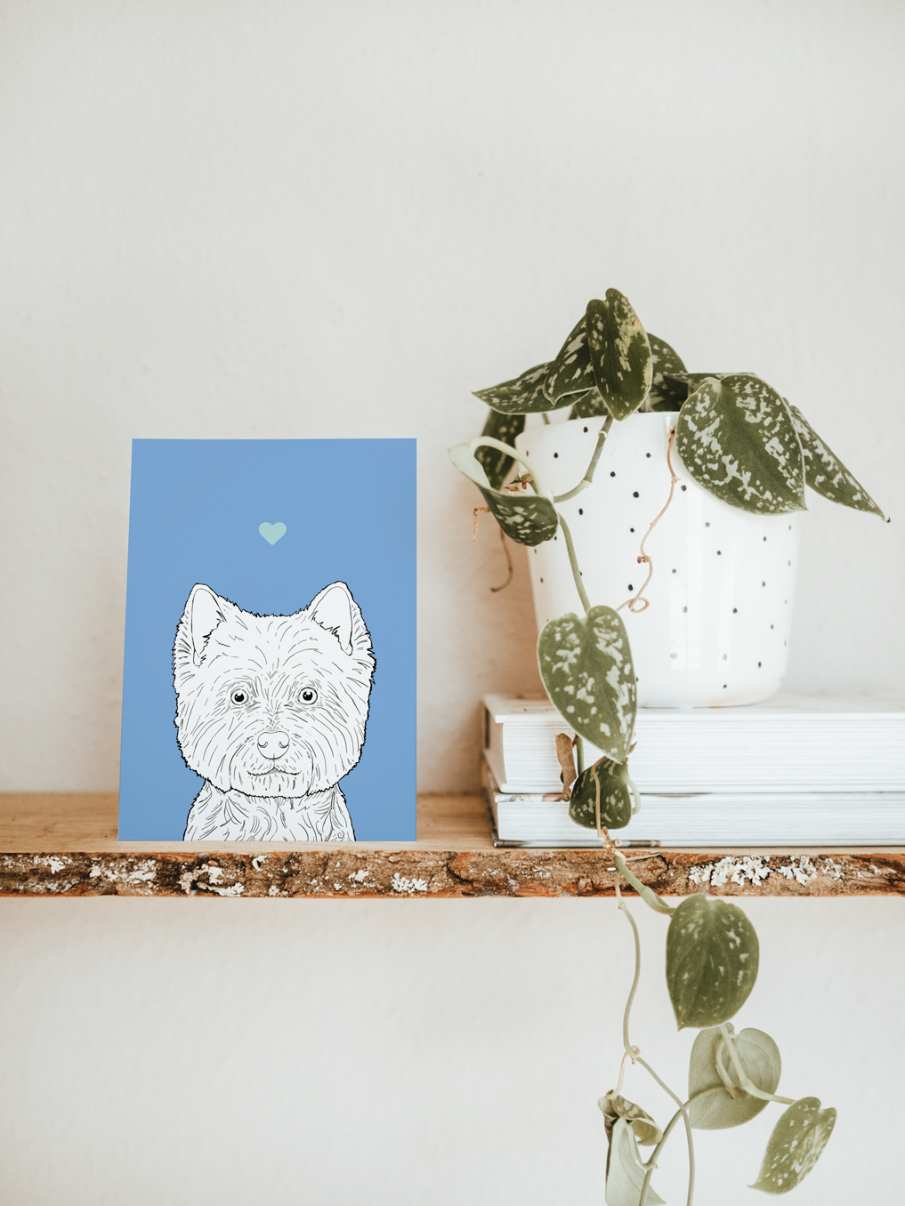 An A6 greetings card & white plant pot on a shelf. The card has a pale blue coloured background, with a black & white digital illustration of the head & shoulders of a Westie dog.