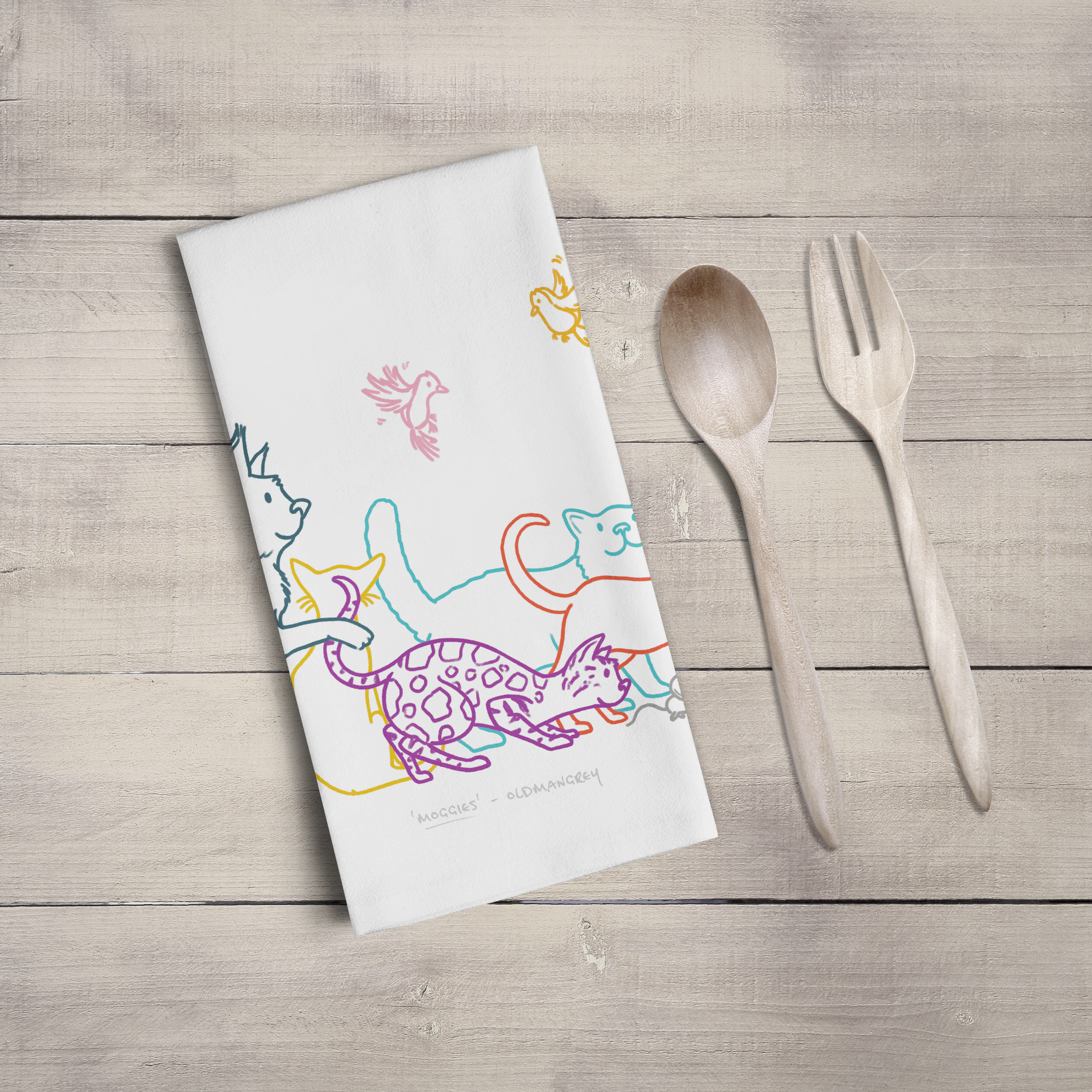 A large tea towel folded to a rectangle on a wooden surface with wooden cutlery. Part of the design can be seen - colourful digitally illustrated cats & butterflies.