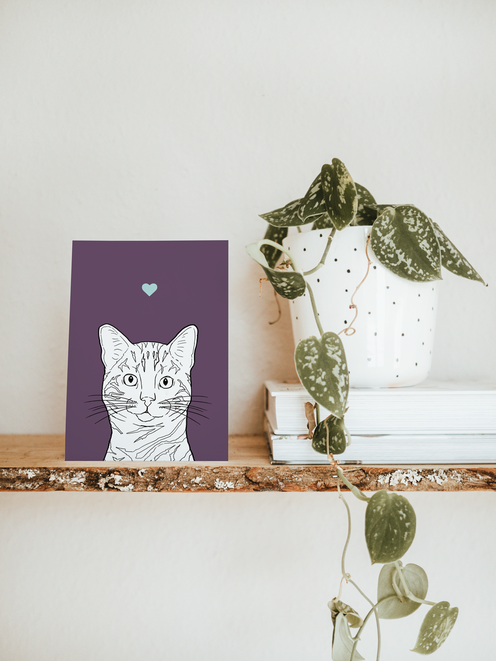 An A6 greetings card & white plant pot on a shelf. The card has a deep purple coloured background, with a black & white digital illustration of the head & shoulders of a Bengal cat.