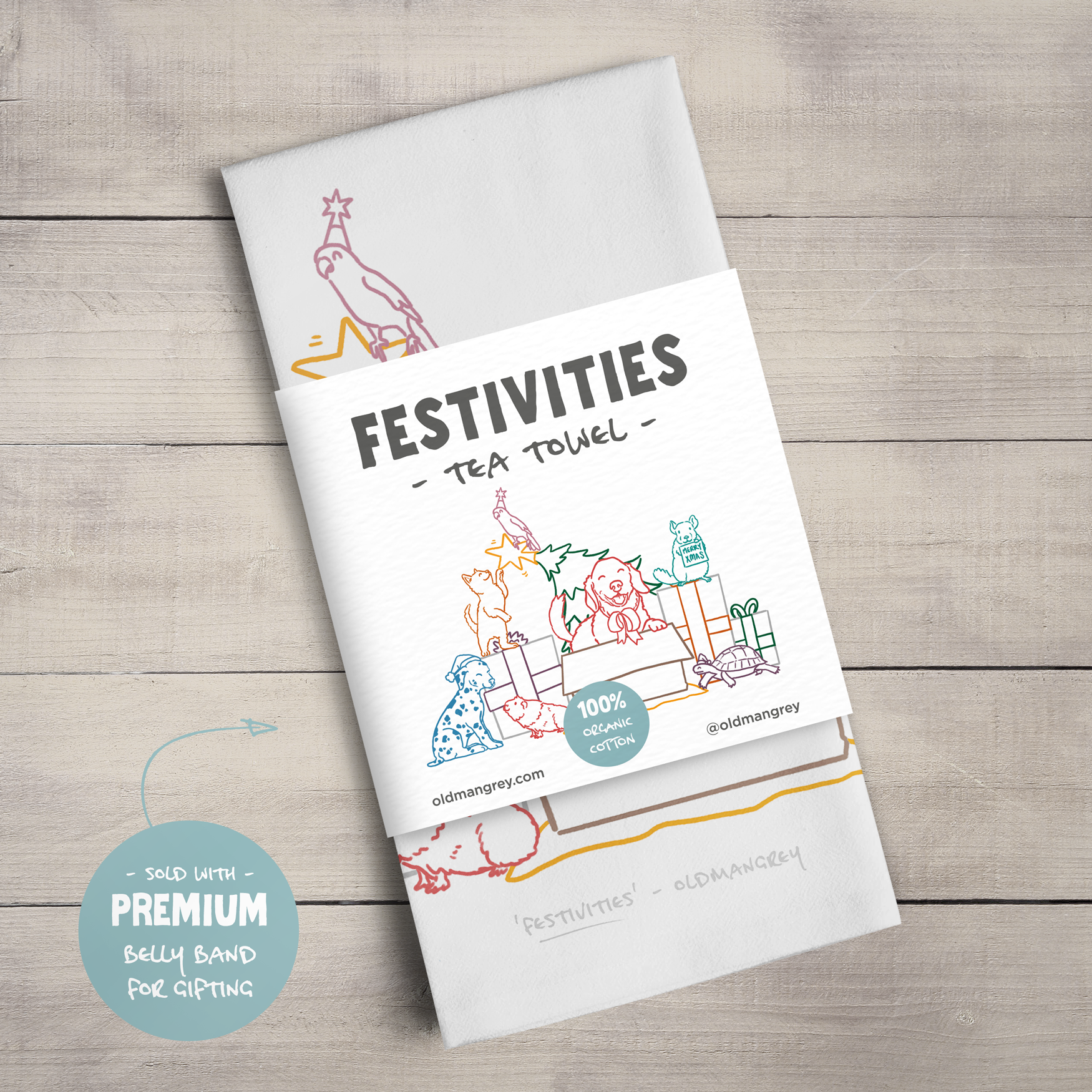 A large tea towel folded to a rectangle wrapped in a paper belly band showing the colourful digital illustration of 7 pets opening presents & playing with the Christmas tree & the title 'Festivities'.