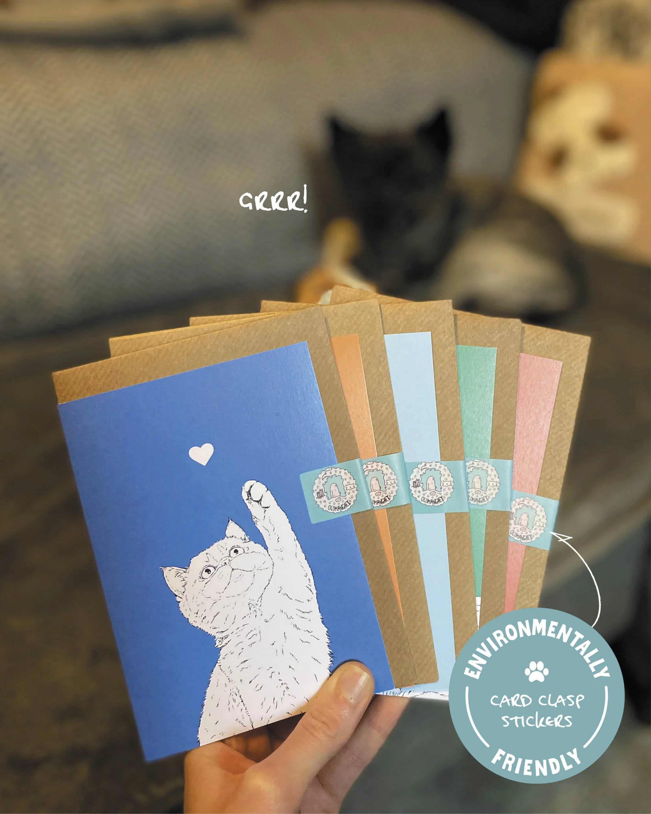 Five A6 cat cards, held up in a fan shape. Each has a brown envelope tucked inside, wrapped in a card clasp sticker. A wee grey dog is in the blurred background, lying on a grey sofa.