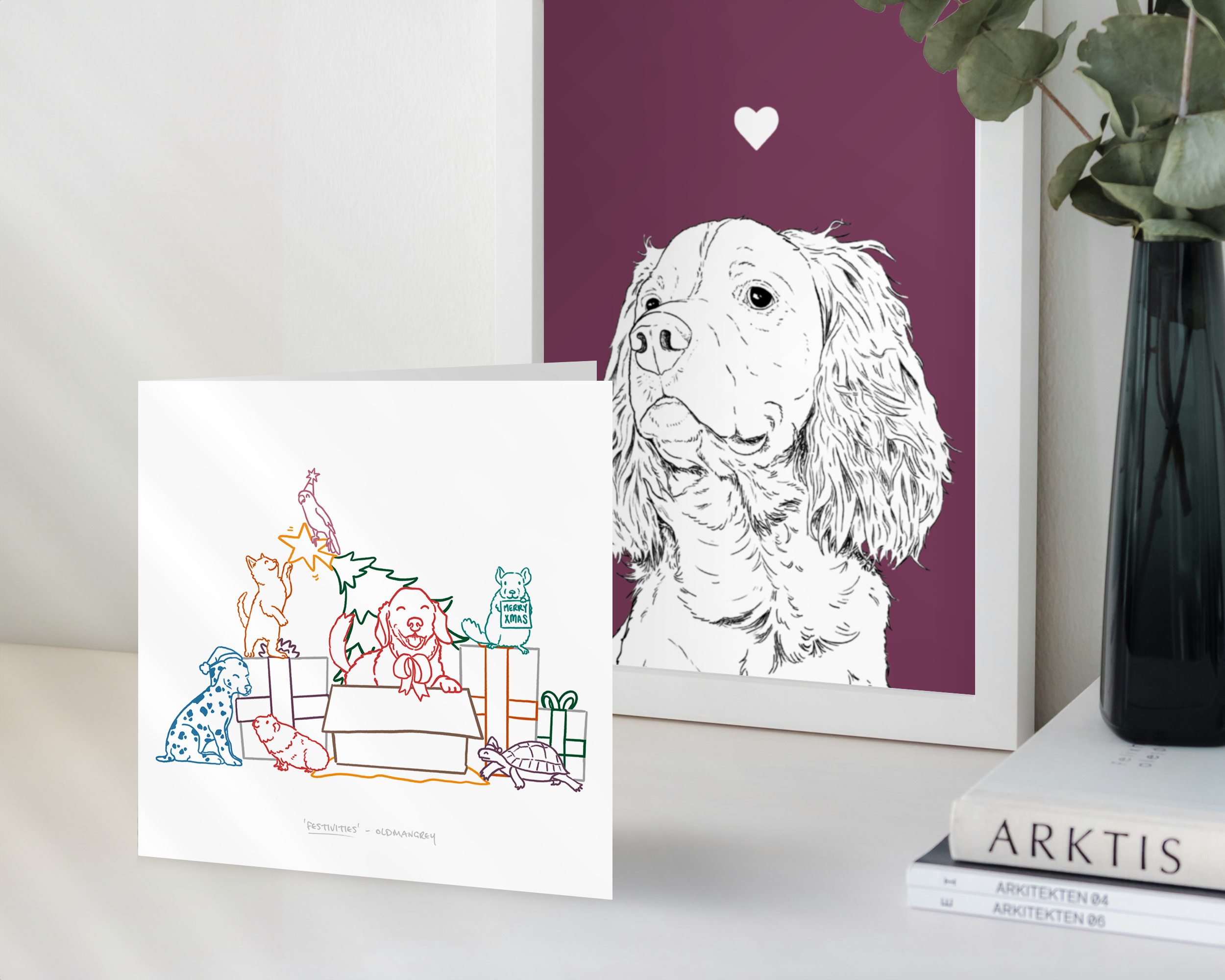 A square Christmas card standing in front of a framed Spaniel dog print. a colourful image of pets including dog, cat, bird & guinea pig opening presents & bending the tree!