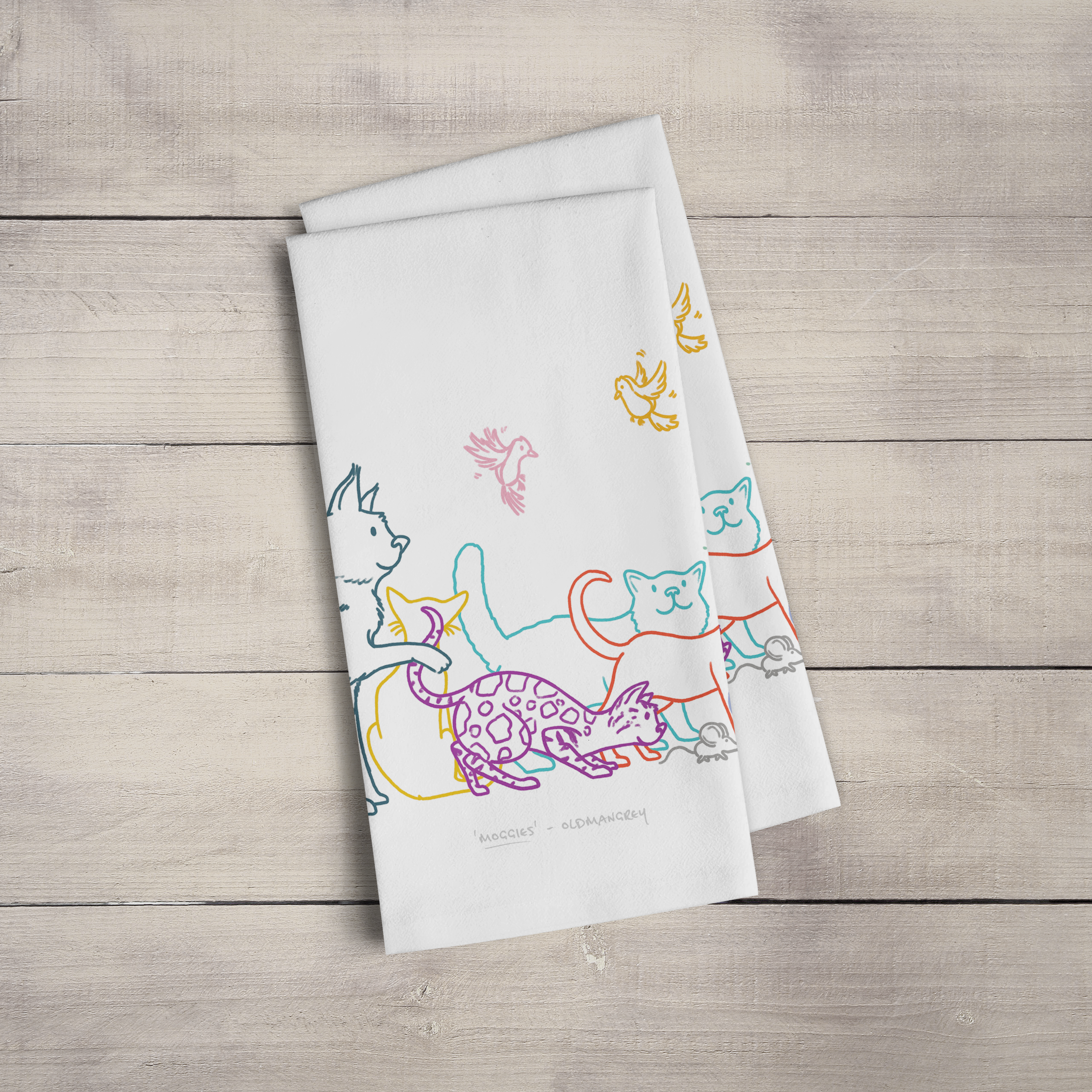 Two large tea towels folded to a rectangle & fanned out, on a wooden surface. Part of the design can be seen - colourful digitally illustrated cats & butterflies.