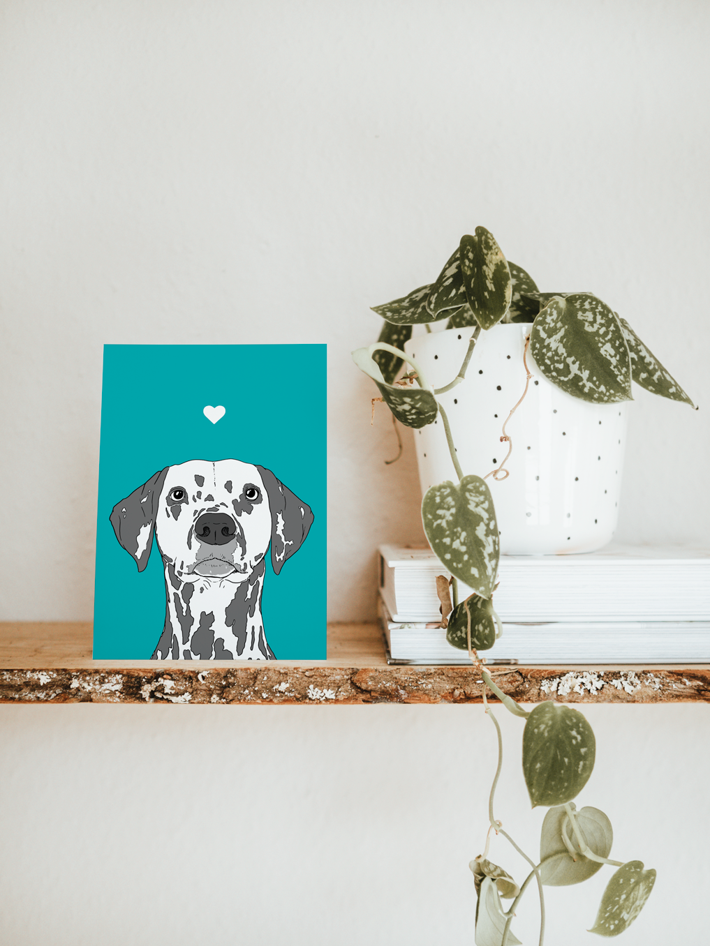 An A6 greetings card & white plant pot on a shelf. The card has a teal blue coloured background, with a black & white digital illustration of the head & shoulders of a Dalmatian dog.