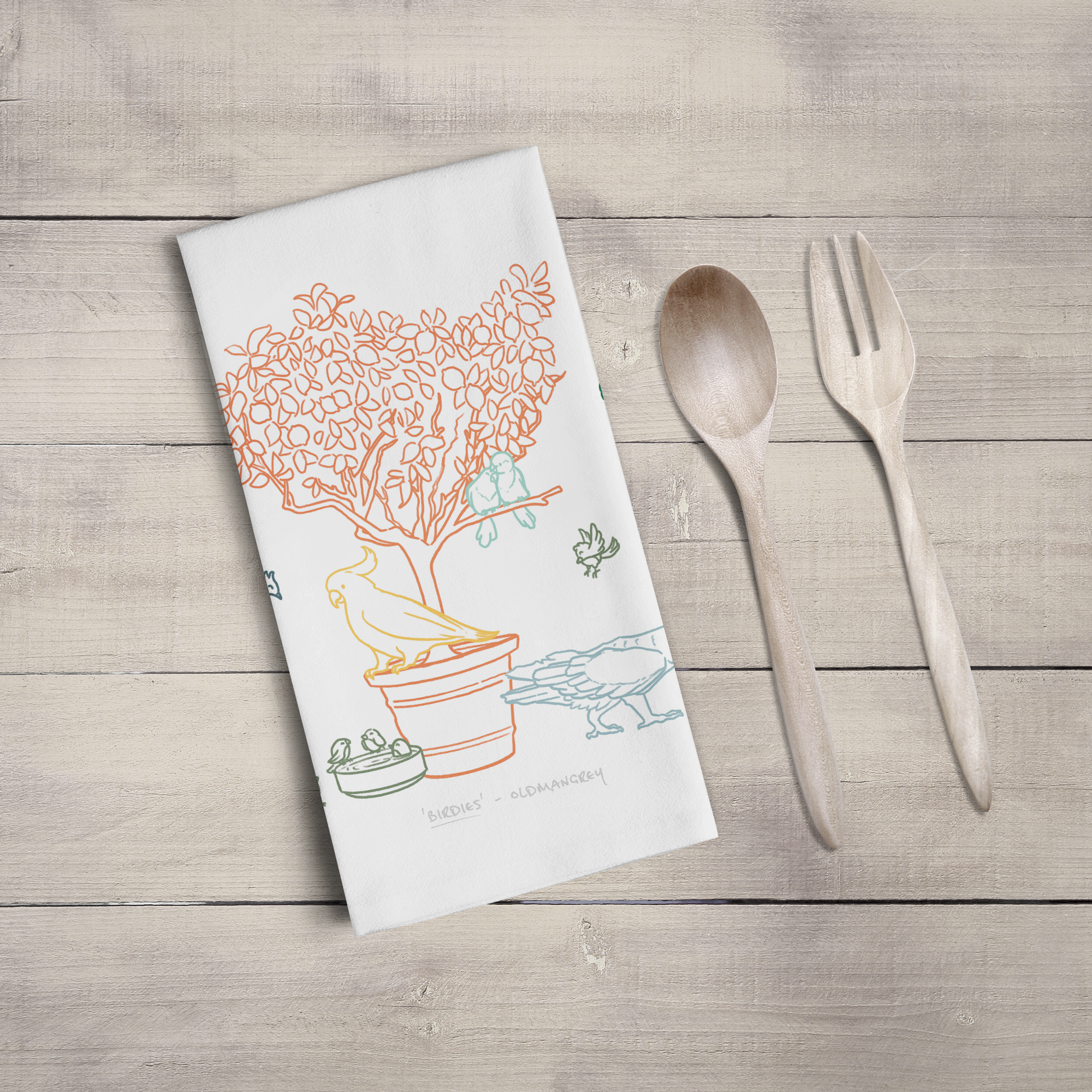 A large tea towel folded to a rectangle on a wooden surface with wooden cutlery. Part of the design can be seen - colourful digitally illustrated birds on & around a small tree.