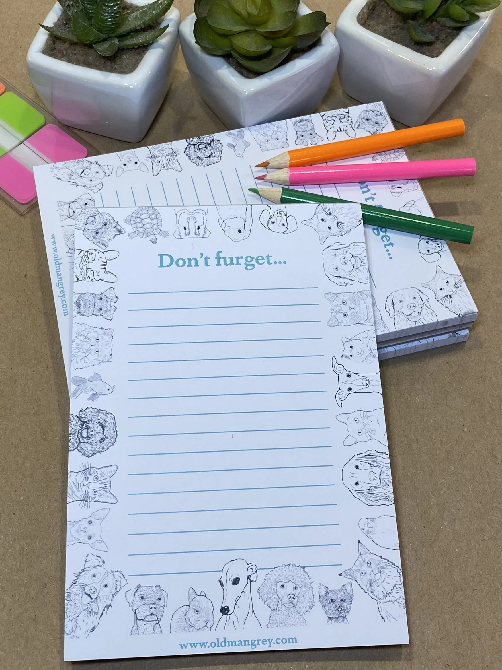 An A6 memo pad titled 'Don't Furget'. It has a black & white border of digitally illustrated pets! Leaning on a pile of other copies, with mini colouring pencils.