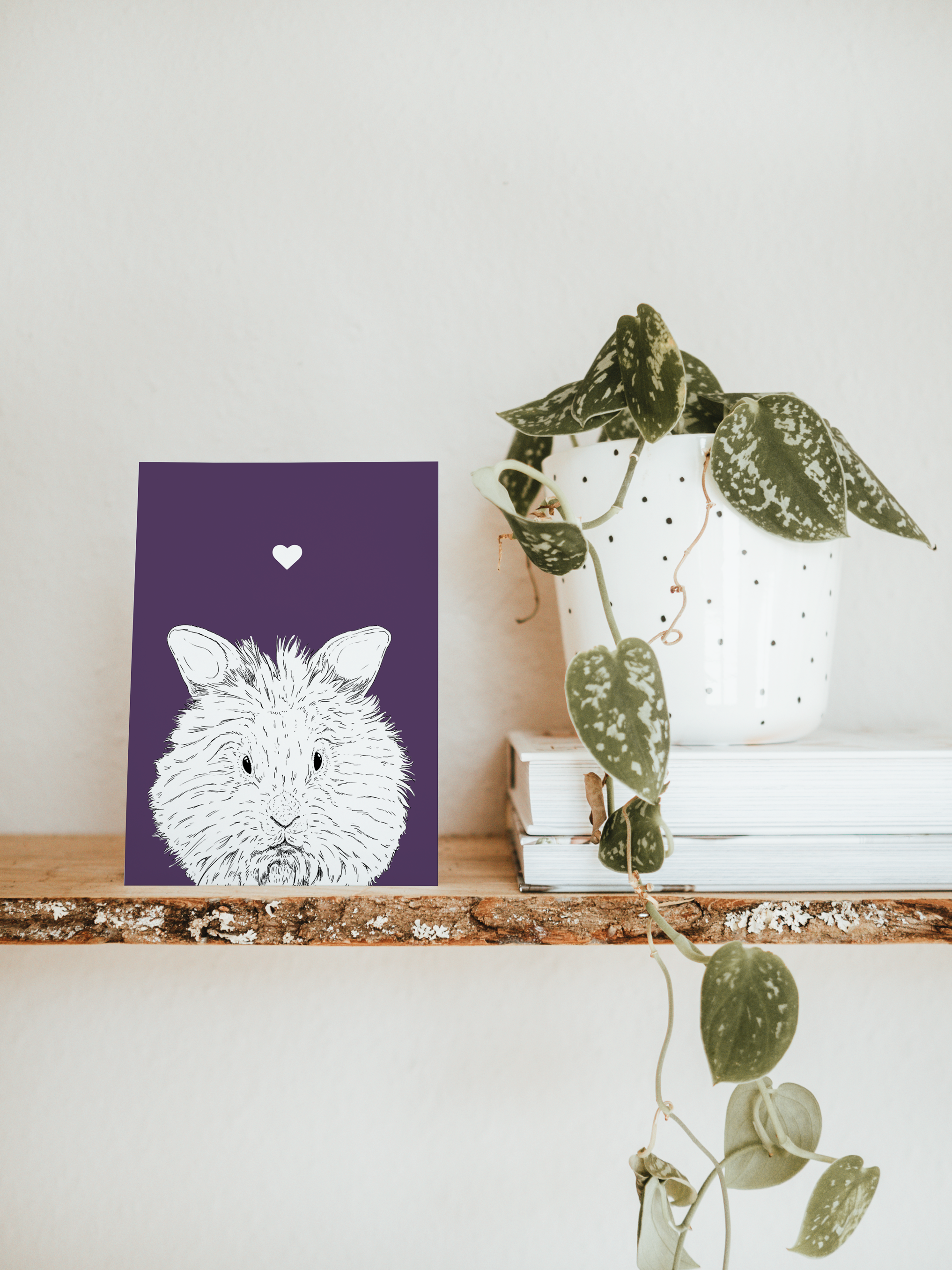 An A6 greetings card & white plant pot on a shelf. The card has a deep purple coloured background, with a black & white digital illustration of the head & shoulders of a furry Lionhead Rabbit.