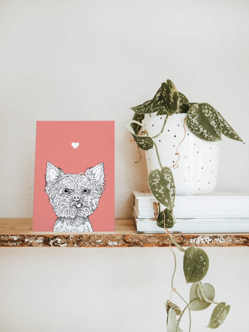 An A6 greetings card & white plant pot on a shelf. The card has a pale pink coloured background, with a black & white digital illustration of the head & shoulders of a Yorkie dog.