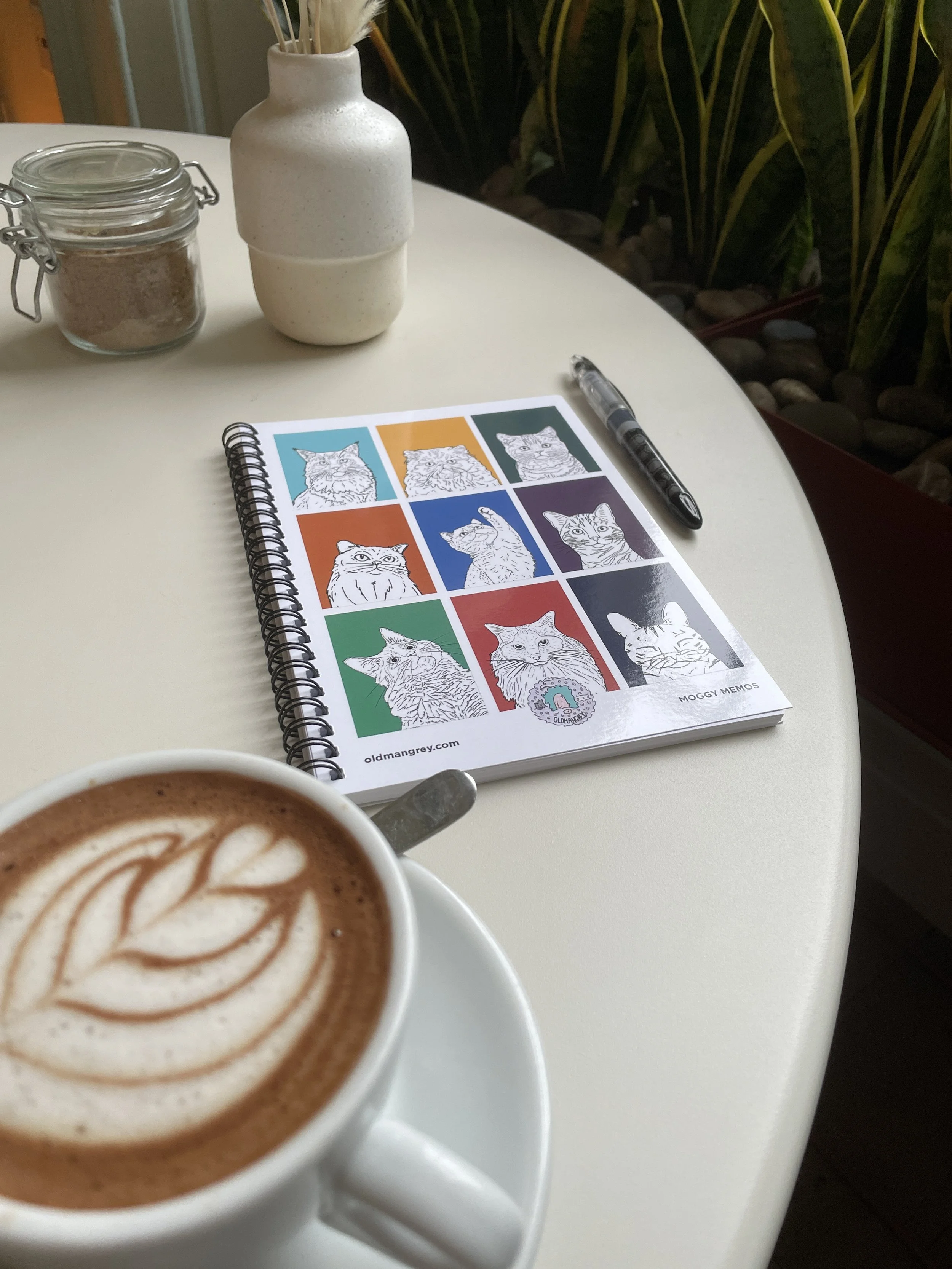 My A5 Moggy Memos wire bound notebook lying on a white table with a coffee & pens next to it. 9 coloured boxes each have a black & white illustration of the head & shoulders of a cat.