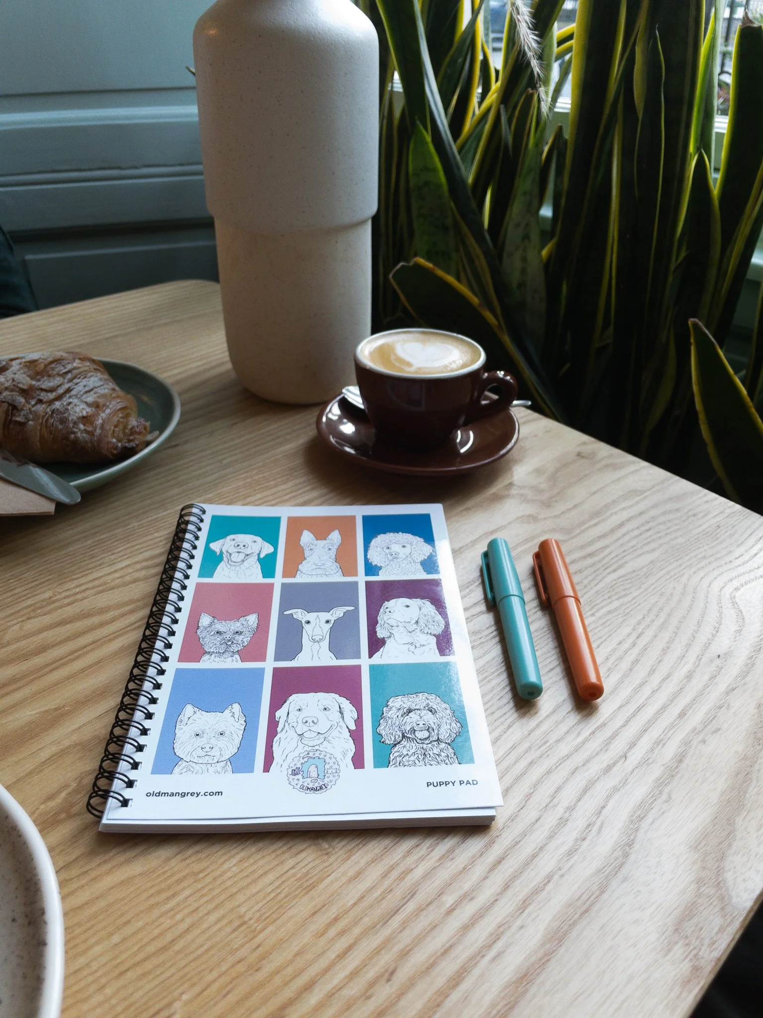 My A5 Puppy Pad wire bound notebook lying on a wooden table with a coffee & plants in the background. 9 coloured boxes each have a black & white illustration of the head & shoulders of a dog.