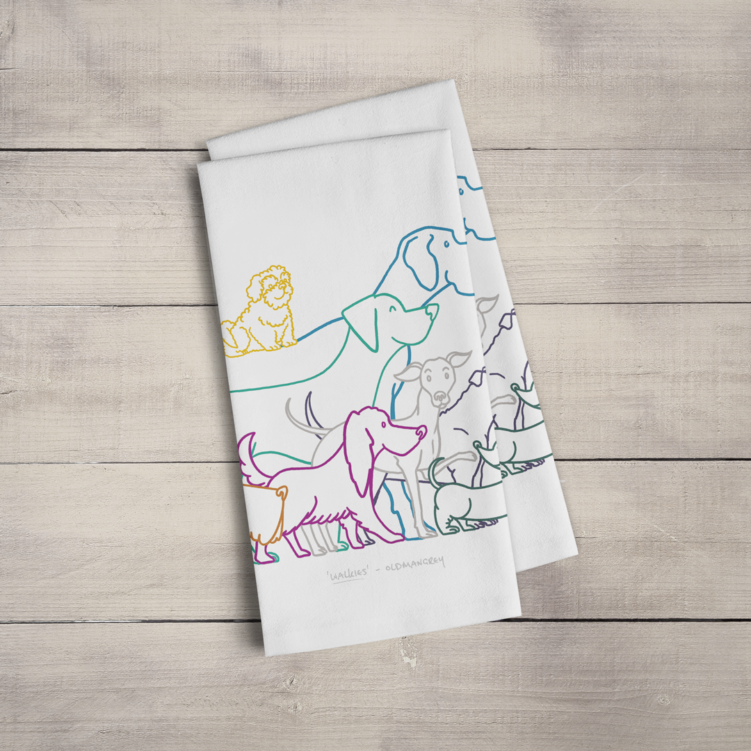 Two large tea towels folded to a rectangle & fanned out, on a wooden surface. Part of the design can be seen - colourful digitally illustrated dogs all eager for walkies.