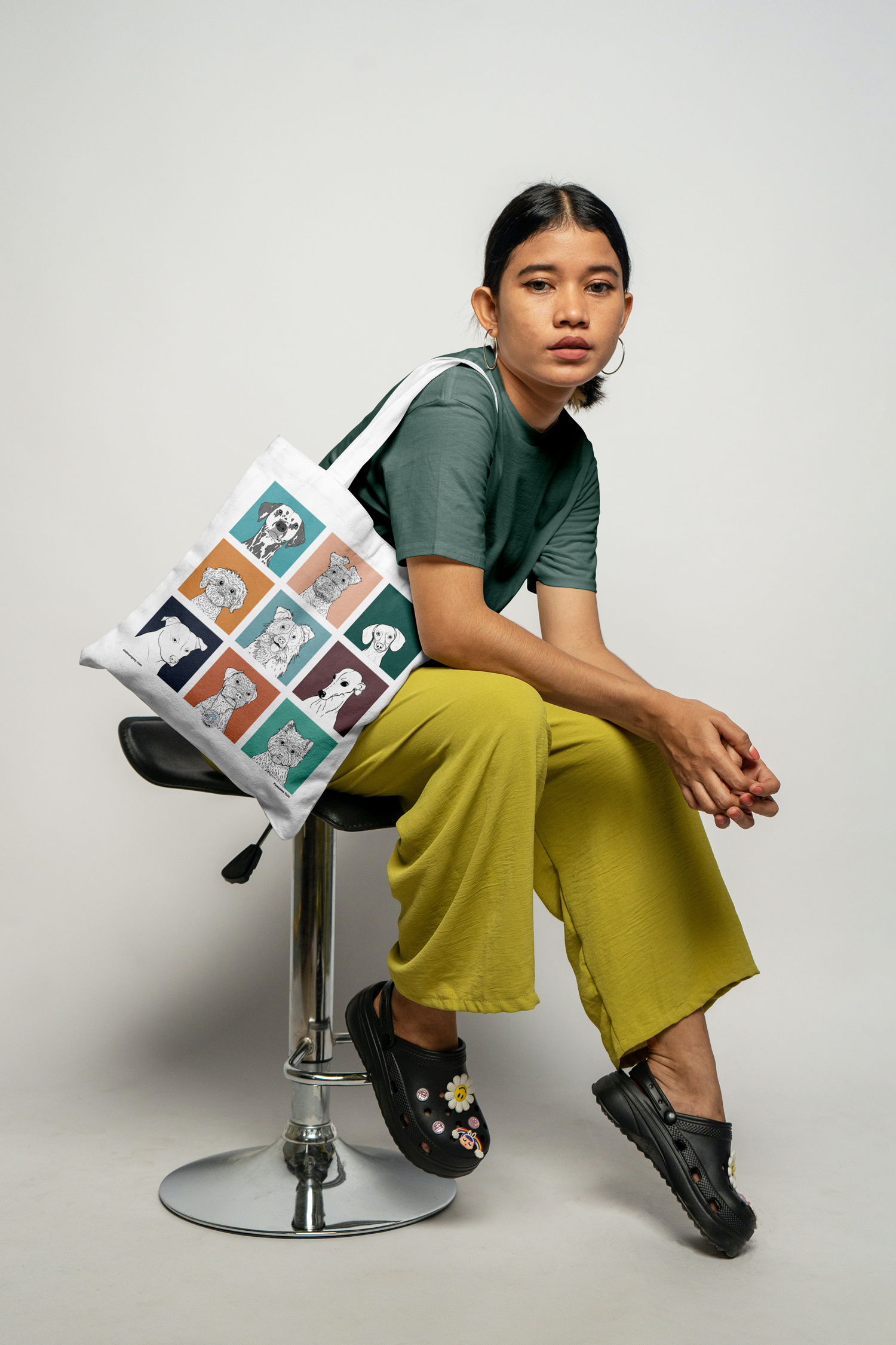 A female model sat sideways on a stool with a tote bag on her shoulder. It has 9 brightly coloured boxes,  with a black & white digital illustration of the head & shoulders of different dog breeds.