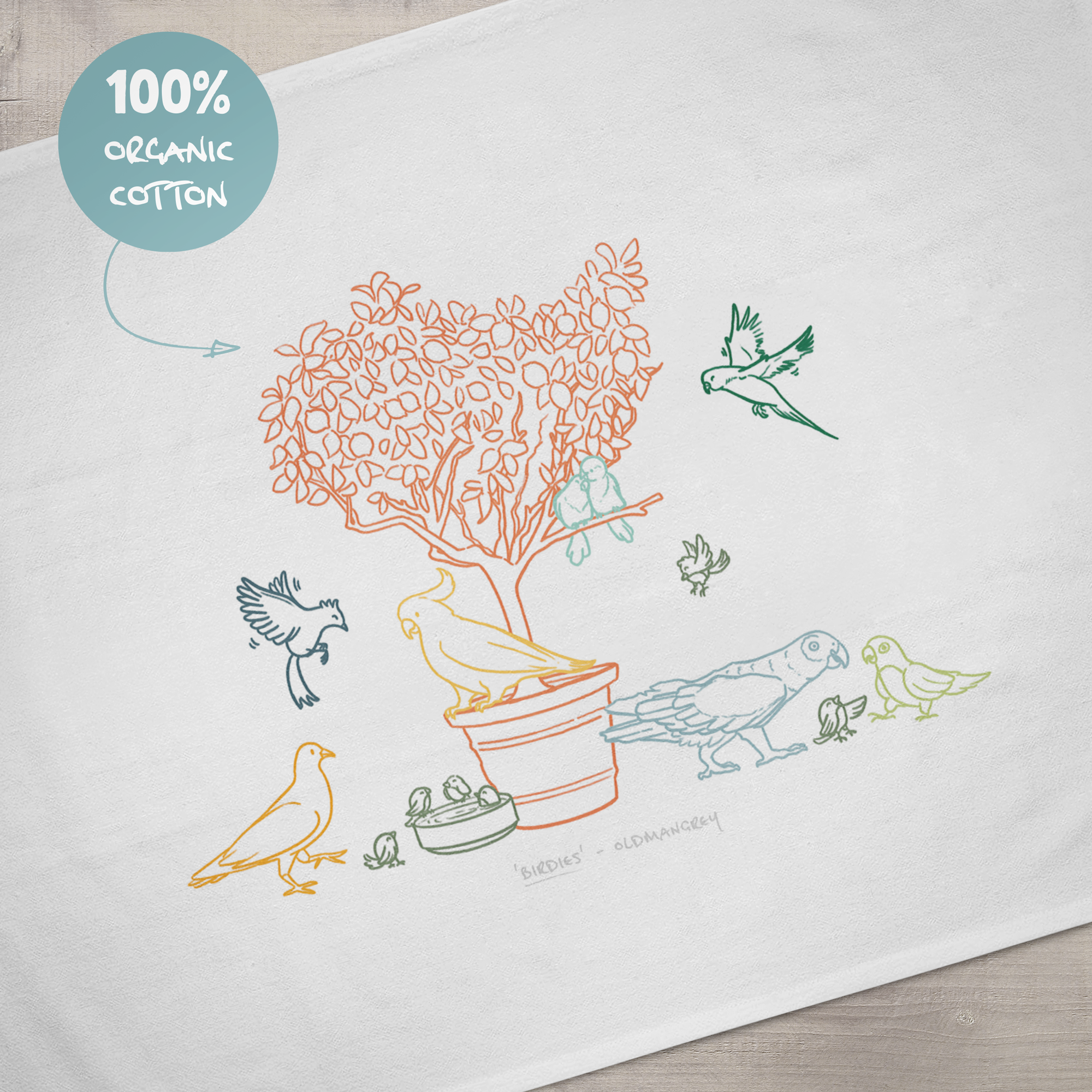 A close up of the design on a large tea towel. It has a colourful digital illustration of birds on & around a small tree. A sign says '100% organic cotton'.