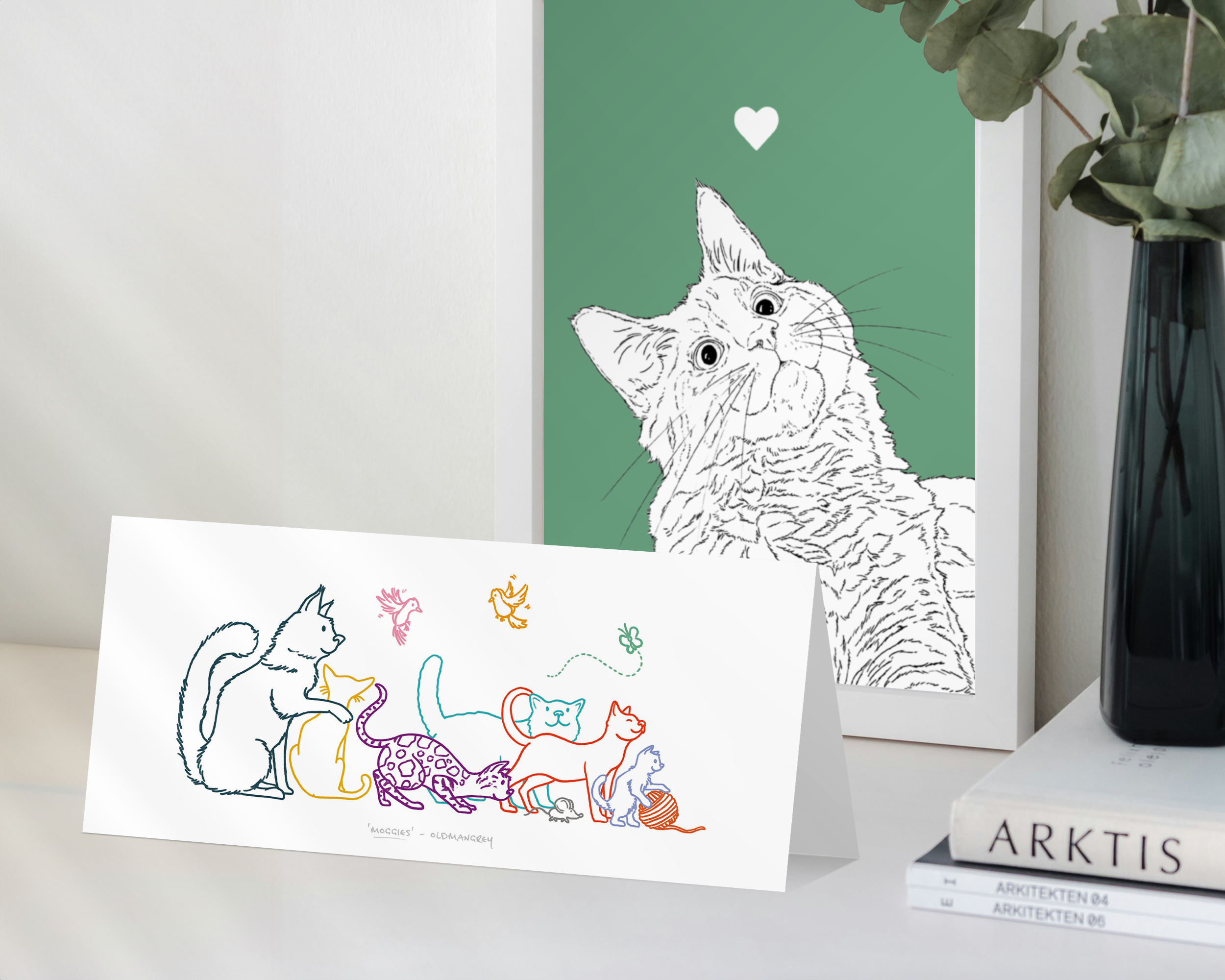 A landscape greetings card standing in front of a framed cat print. The card has a colourful digital illustration of 6 cats, 2 birds, a butterfly & mouse.