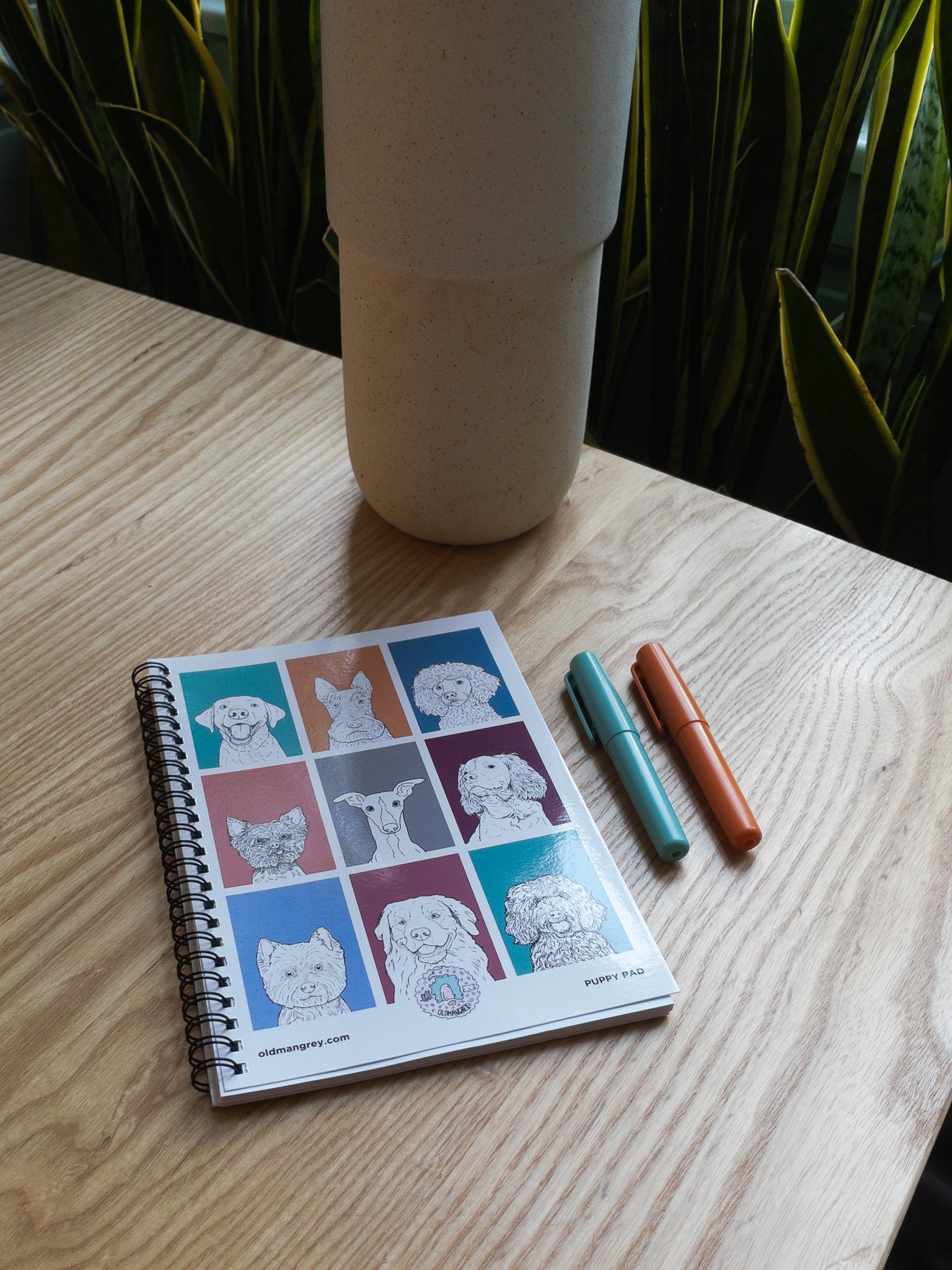 My A5 Puppy Pad wire bound notebook lying on a wooden table with a coffee, pastry & pens. 9 coloured boxes each have a black & white illustration of the head & shoulders of a dog.