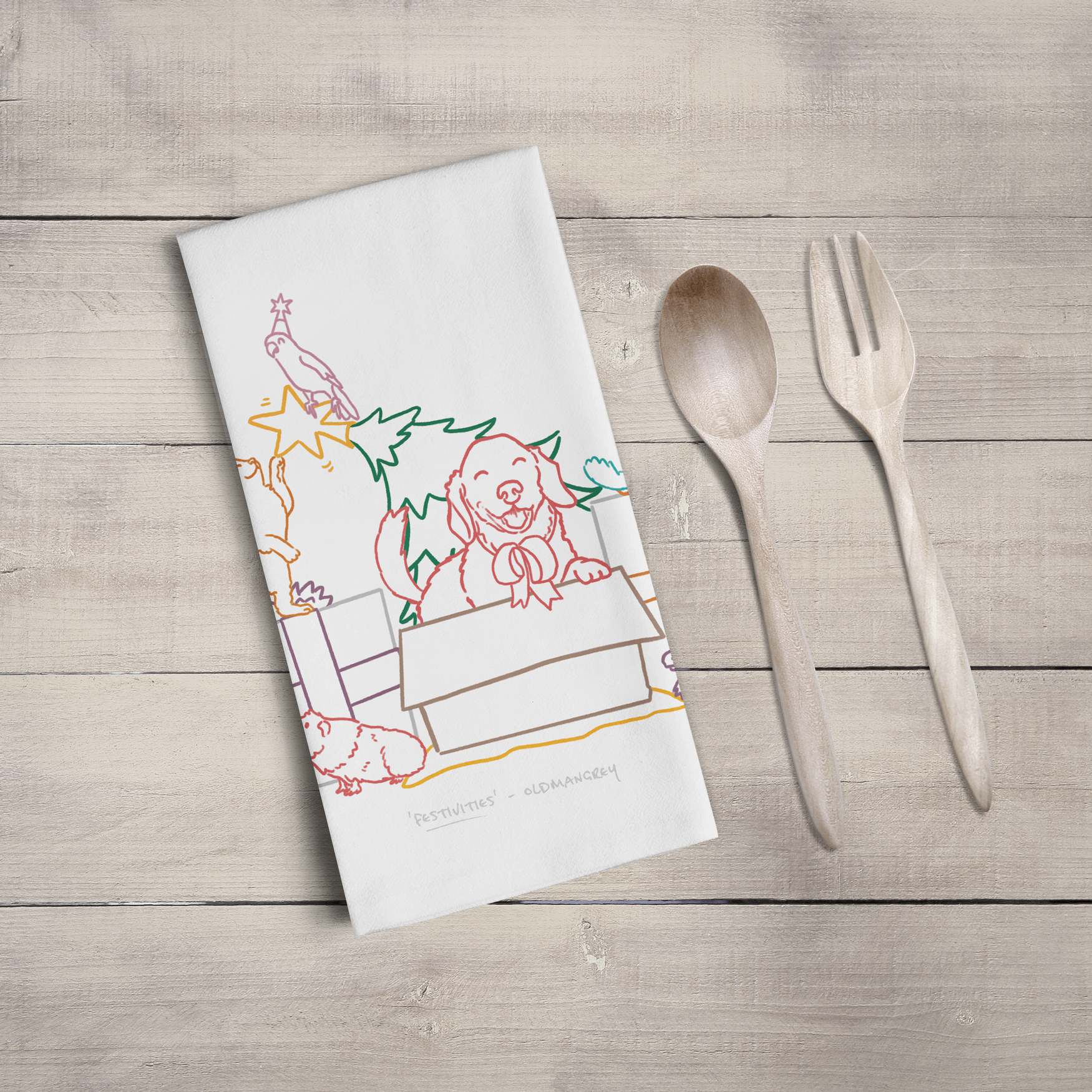 A large tea towel folded to a rectangle on a wooden surface with wooden cutlery. Part of the design can be seen - colourful digitally illustrated pets on & around Christmas gifts & tree.