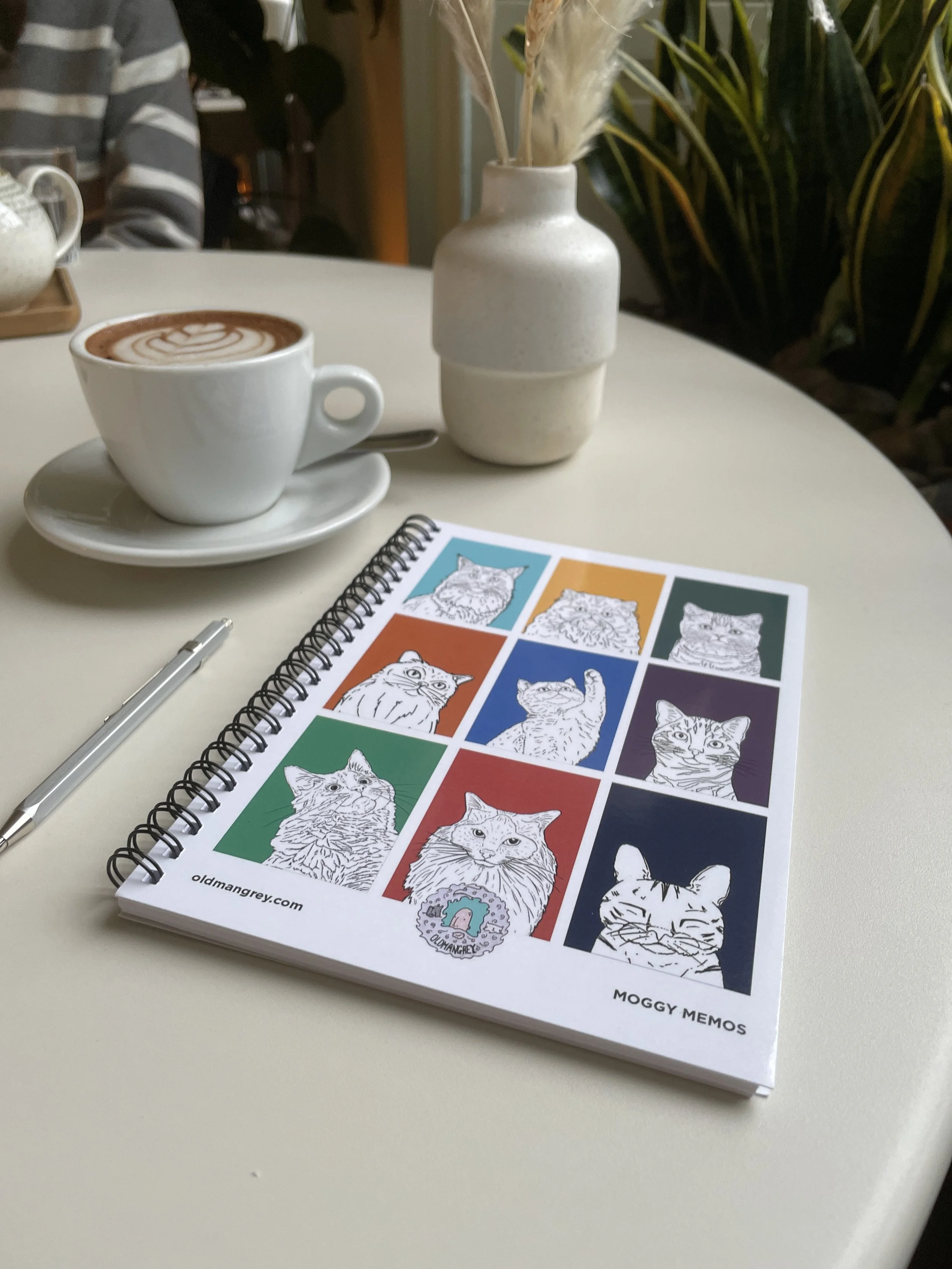My A5 Moggy Memos wire bound notebook lying on a white table with a coffee & plants in the background. 9 coloured boxes each have a black & white illustration of the head & shoulders of a cat.
