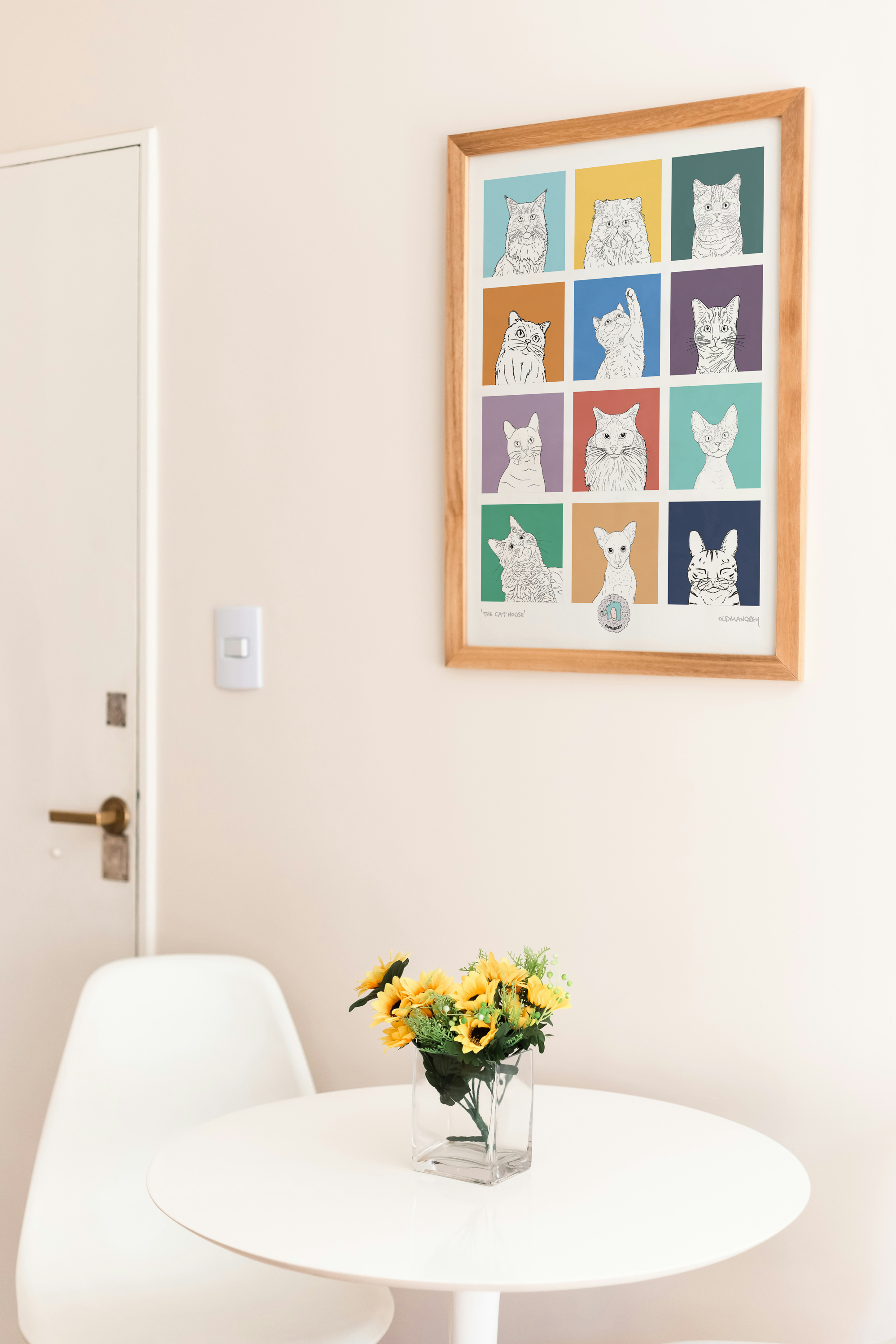 An A3 print with a 3 by 3 grid of bright squares with a black & white digital illustration of the head & shoulders of a cat. Hung above a round white table with yellow flowers.