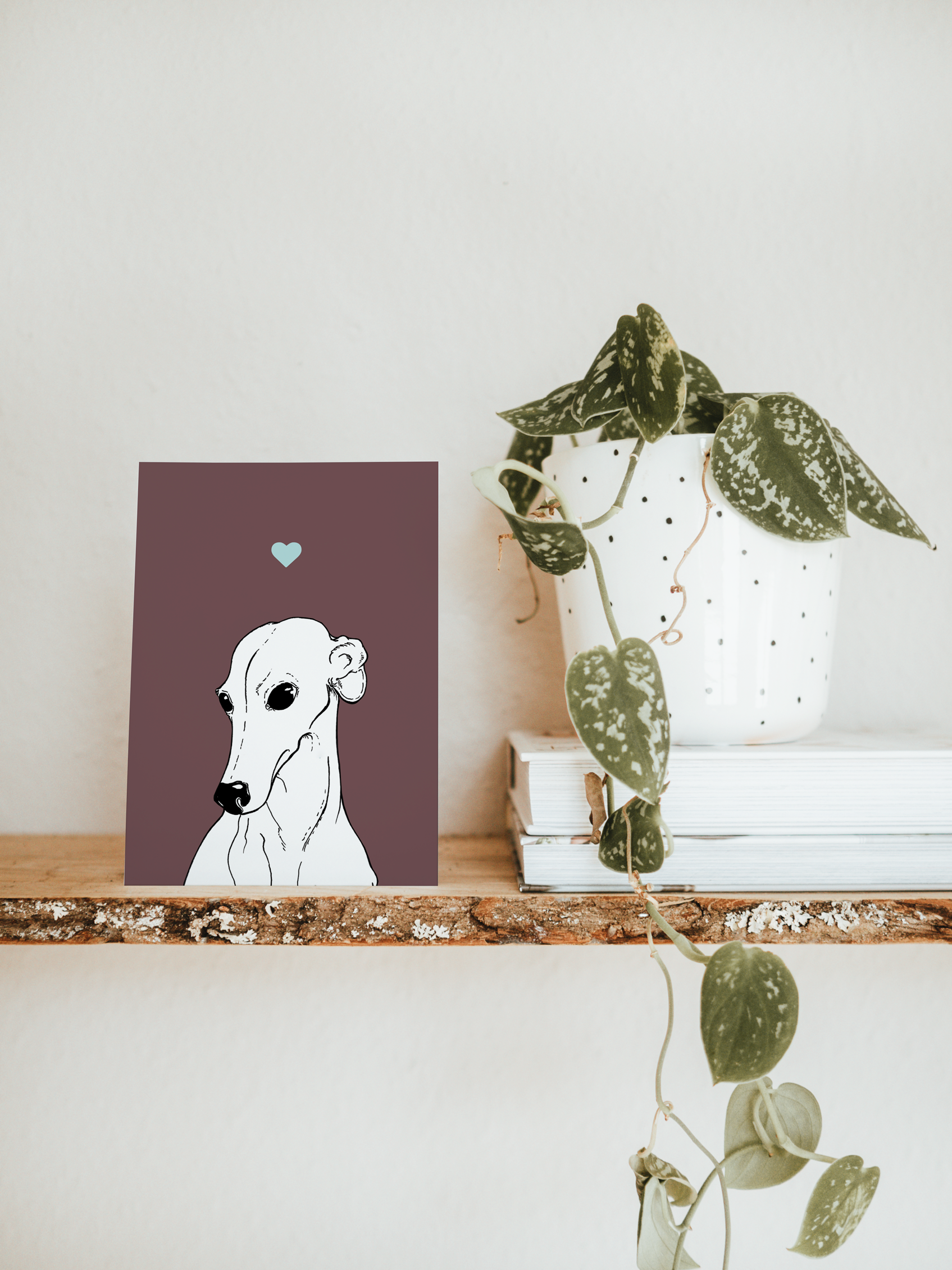 An A6 greetings card & white plant pot on a shelf. The card has a deep purple coloured background, with a black & white digital illustration of the head & shoulders of a Greyhound dog.