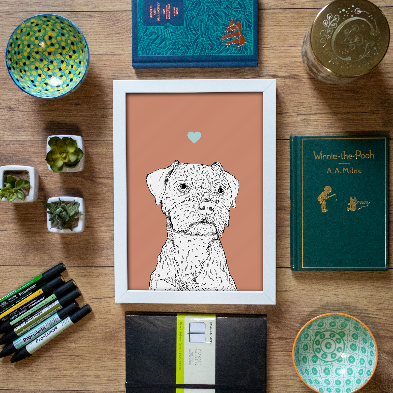An A4 print in a white frame in a flatlay with stationery & home accessories. It has a black & white illustration of the head & shoulders of a Border Terrier dog on a pink red background.
