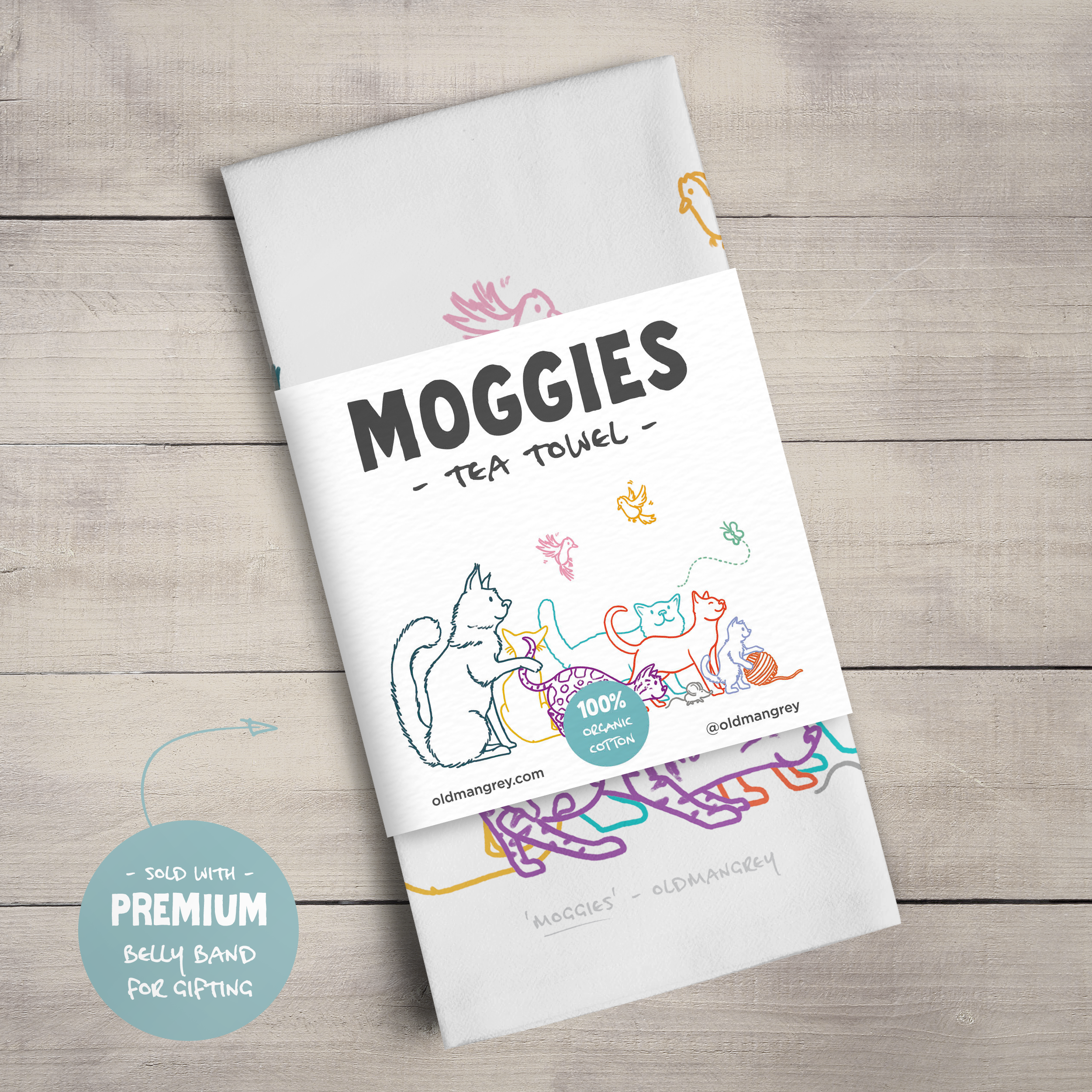 A large tea towel folded to a rectangle wrapped in a paper belly band. This shows the colourful digital illustration of 6 cats, 2 birds, a butterfly, & mouse and the title 'Moggies'.