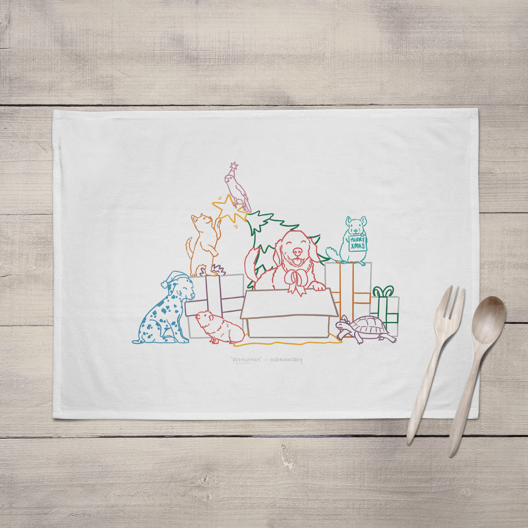 A large tea towel lying open on a grey brown wooden surface with wooden cutlery. It has a colourful digital illustration of 7 pets opening Christmas presents & playing with the Christmas tree!