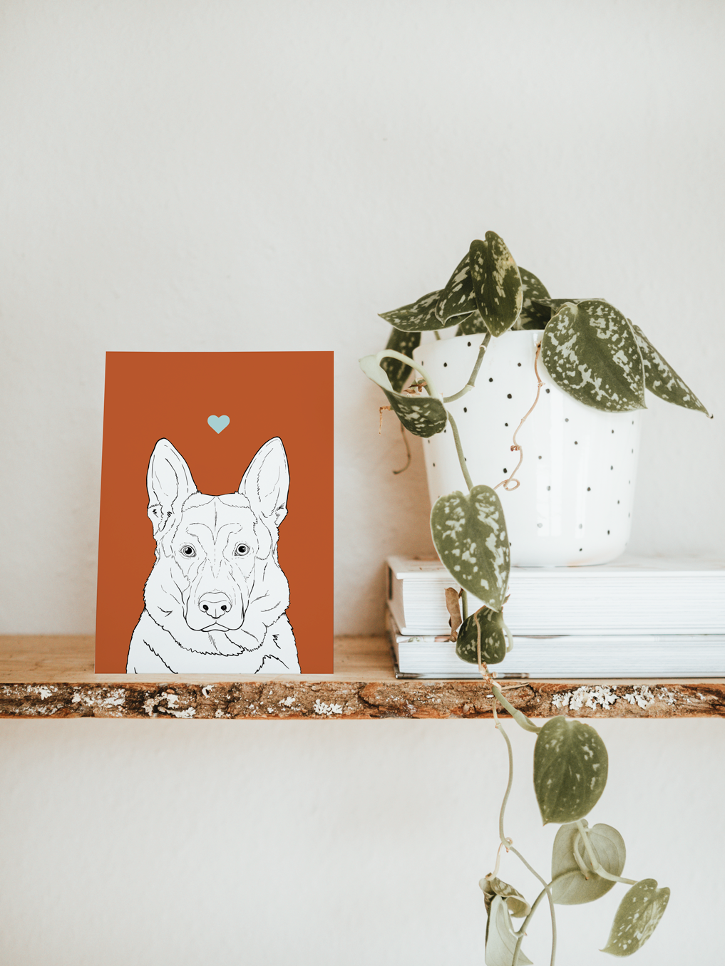 An A6 greetings card & white plant pot on a shelf. The card has a deep red coloured background, with a black & white digital illustration of the head & shoulders of an Alsatian / German Shepherd dog.