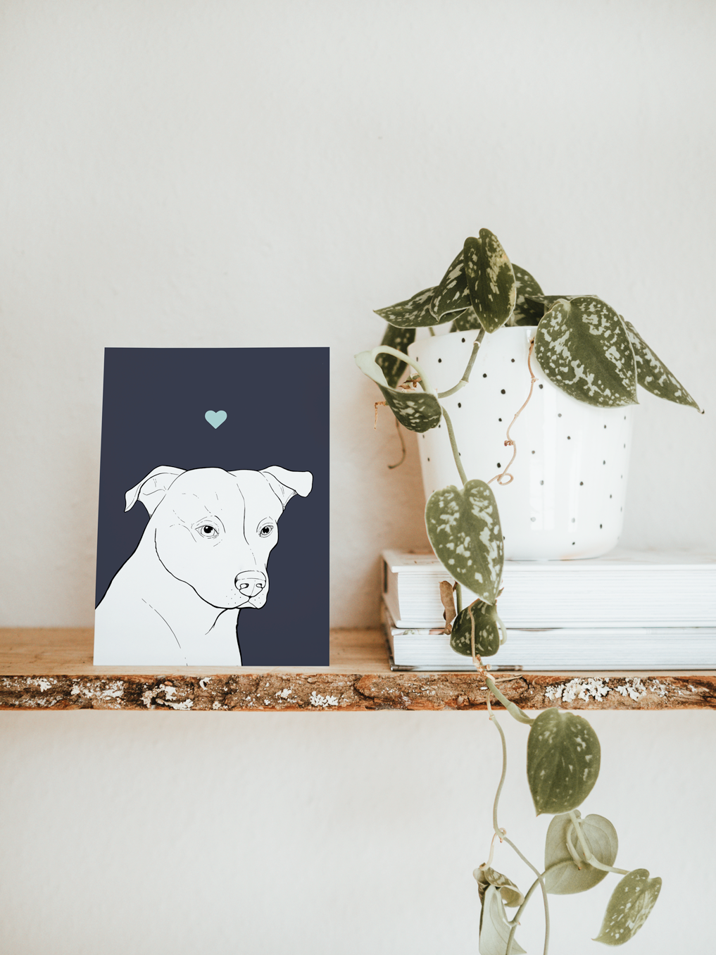 An A6 greetings card & white plant pot on a shelf. The card has a navy blue coloured background, with a black & white digital illustration of the head & shoulders of a Staffie dog.