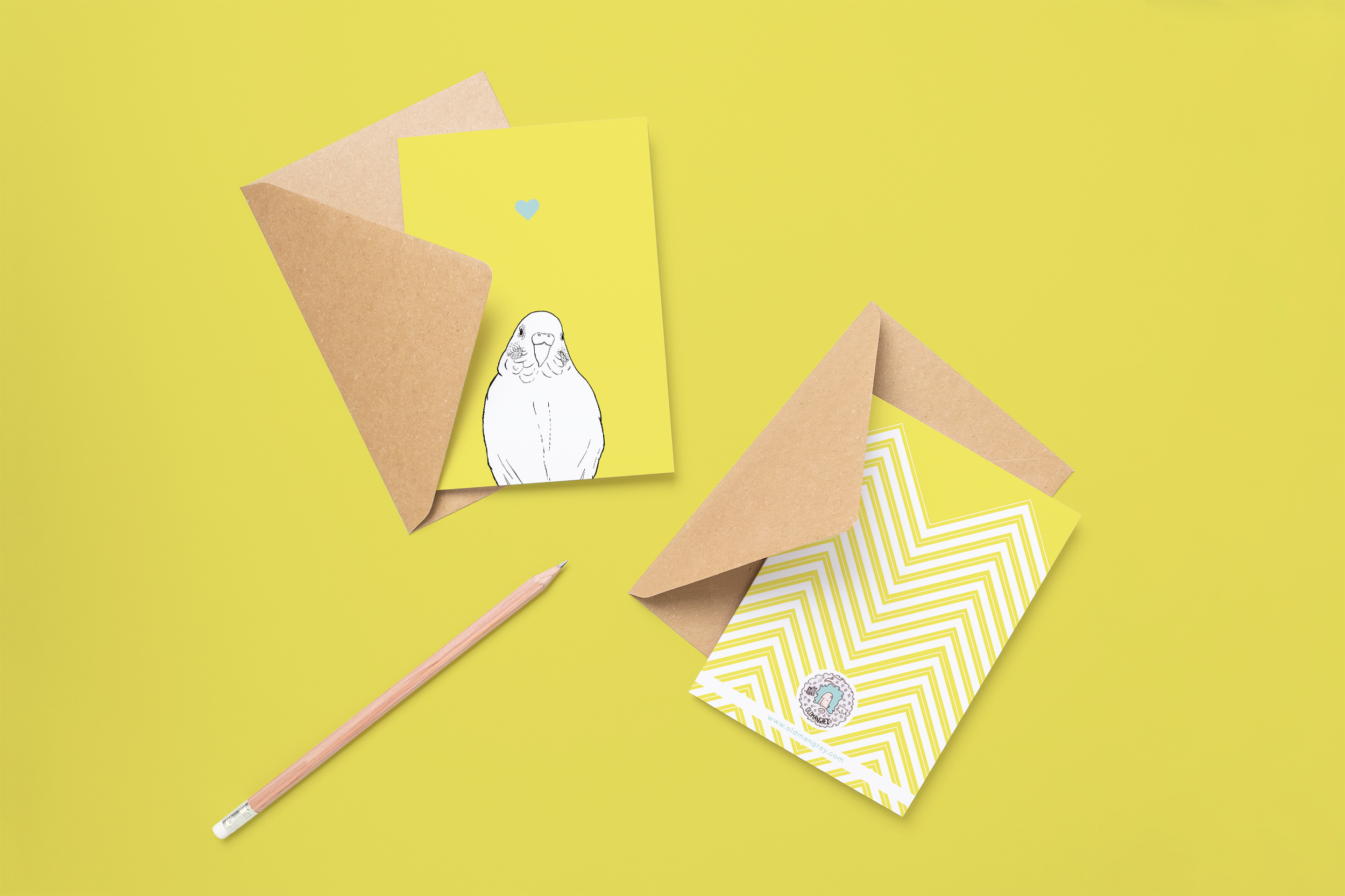 Two cards on brown envelopes, one face up one down. The card and image have a yellow coloured background, with a black & white digital illustration of the head & shoulders of a budgie.