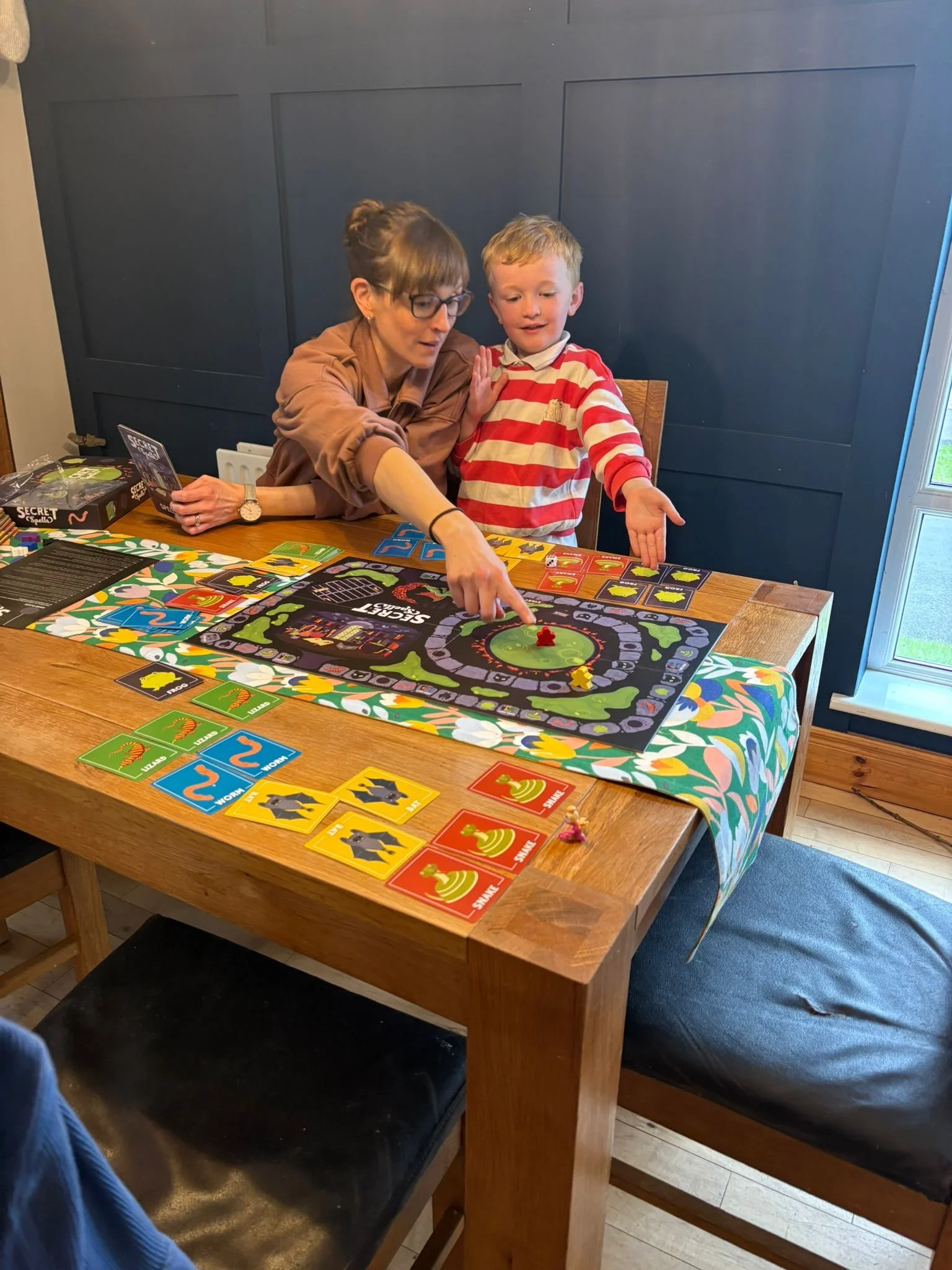 A photo of a woman and a five year old boy playing Secret Spells Boardgame at a dining table. The boy is kneeling up on the chair, holding out his hand excitedly at the board and the woman is pointing at it. Designed by Graeme A Clark aka Oldmangrey.