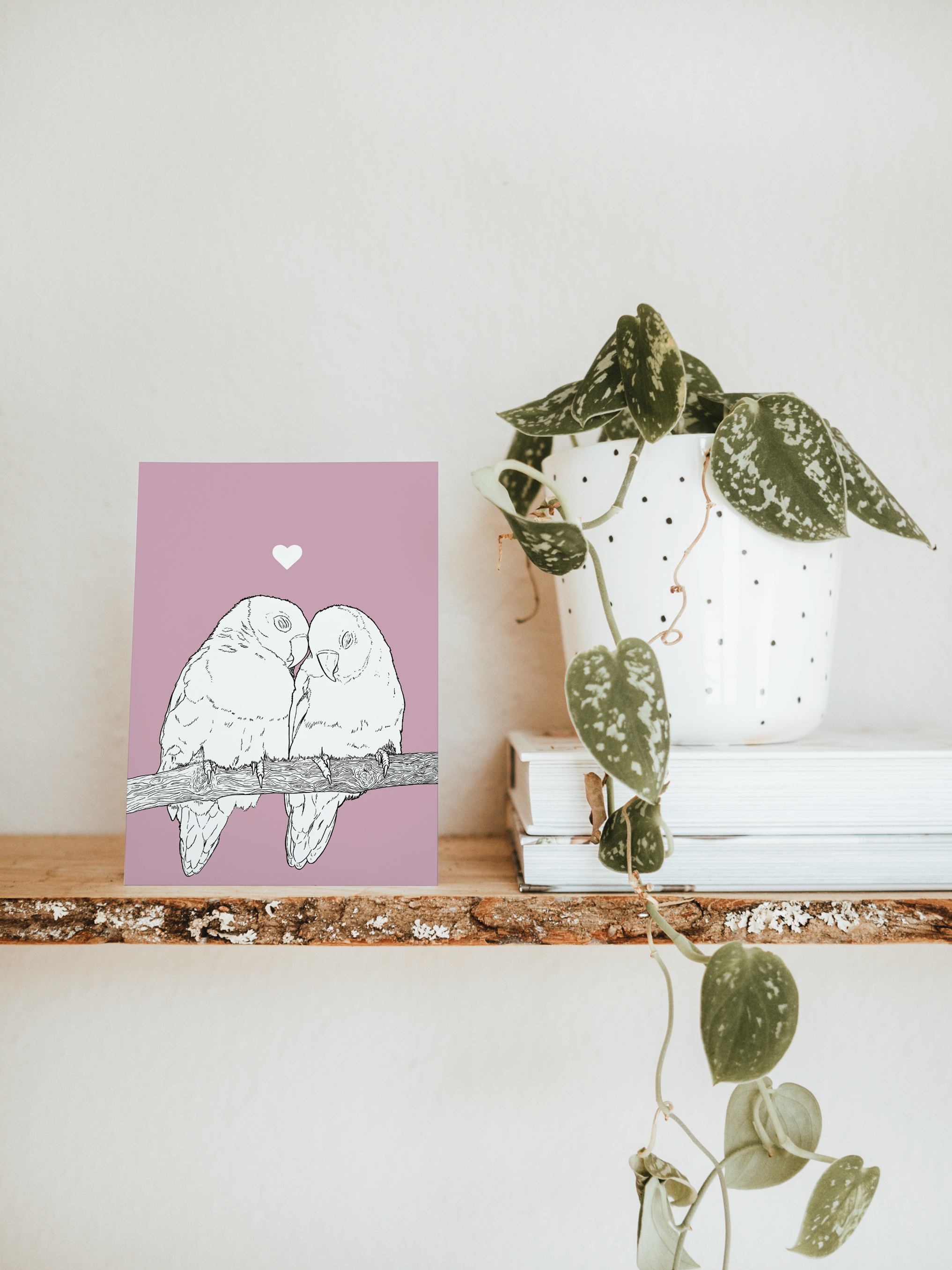 An A6 greetings card & white plant pot on a shelf. The card has a lilac pink coloured background, with a black & white digital illustration of two Lovebirds sat on a branch.