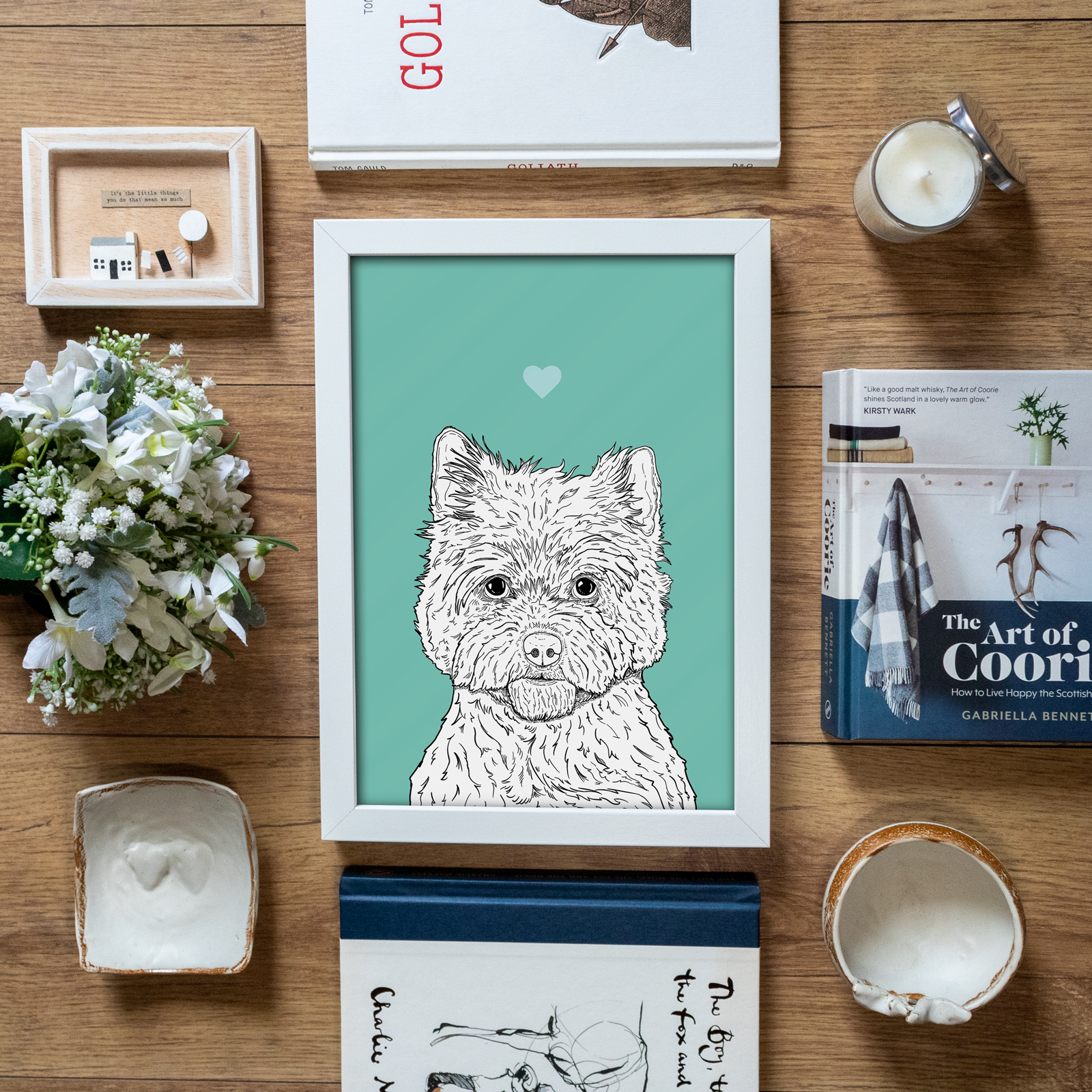 An A4 print in a white frame in a flatlay with stationery & home accessories. It has a black & white illustration of the head & shoulders of a Cairn Terrier dog on a mint green background.