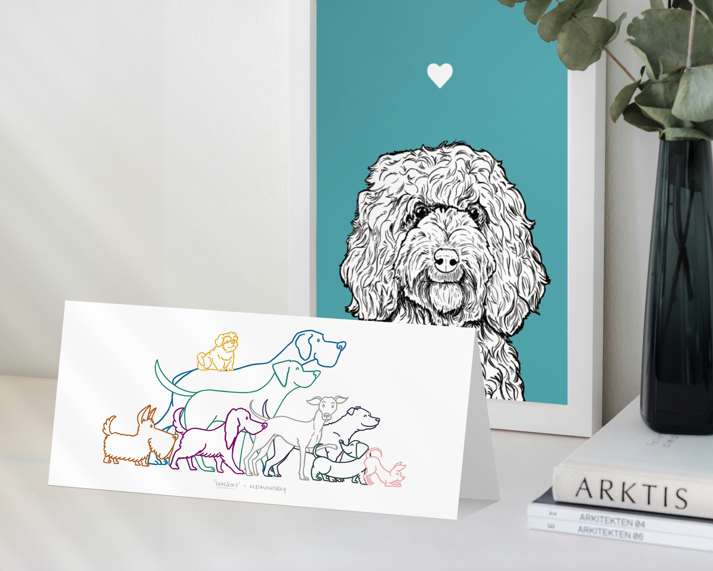 A landscape greetings card standing in front of a framed Cockapoo dog print. The card has a colourful digital illustration of 9 dogs eager to go walkies.