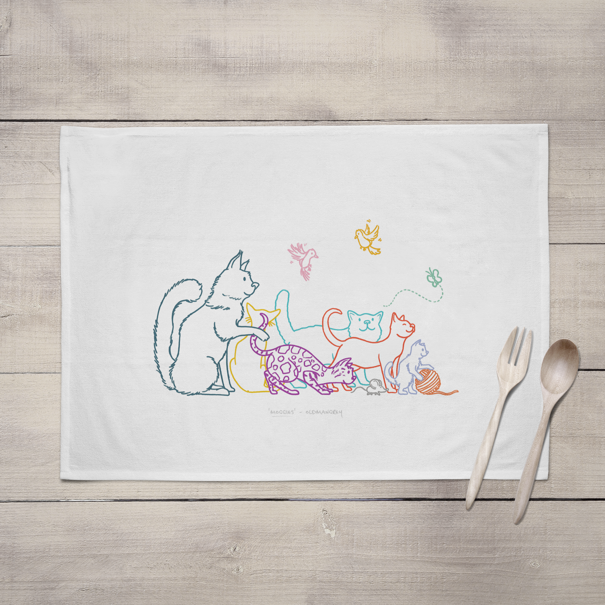 A large tea towel lying open on a grey brown wooden surface with wooden cutlery. It has a colourful digital illustration of 6 cats, 2 birds, a butterfly & mouse.