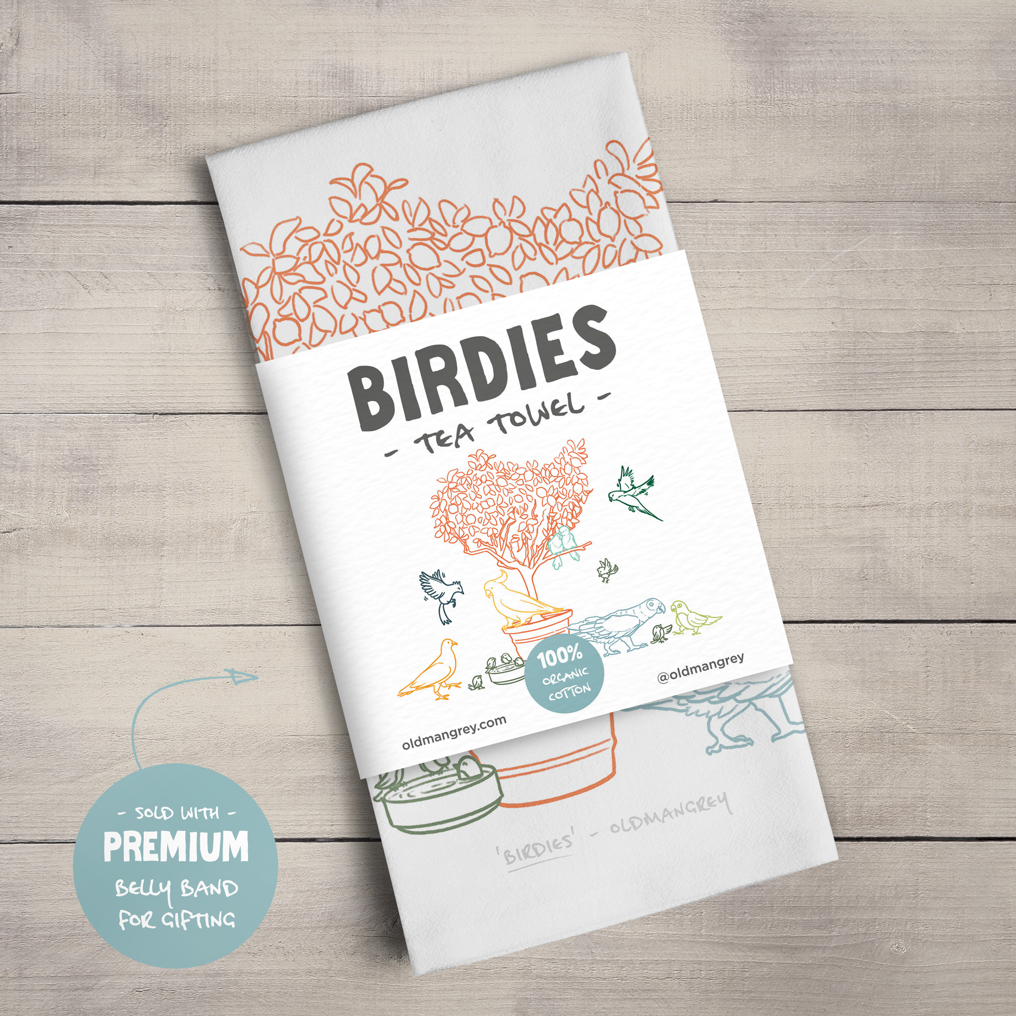 A large tea towel folded to a rectangle wrapped in a paper belly band. This shows the colourful digital illustration of birds on & around a small tree & the title 'Birdies'.
