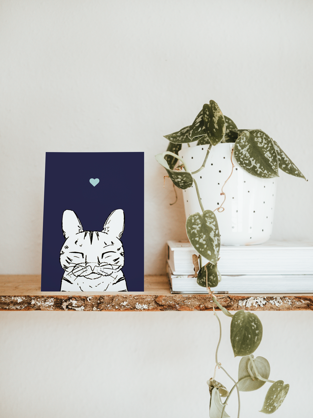 An A6 greetings card & white plant pot on a shelf. The card has a navy blue coloured background, with a black & white digital illustration of the head & shoulders of a stripy sleeping cat.