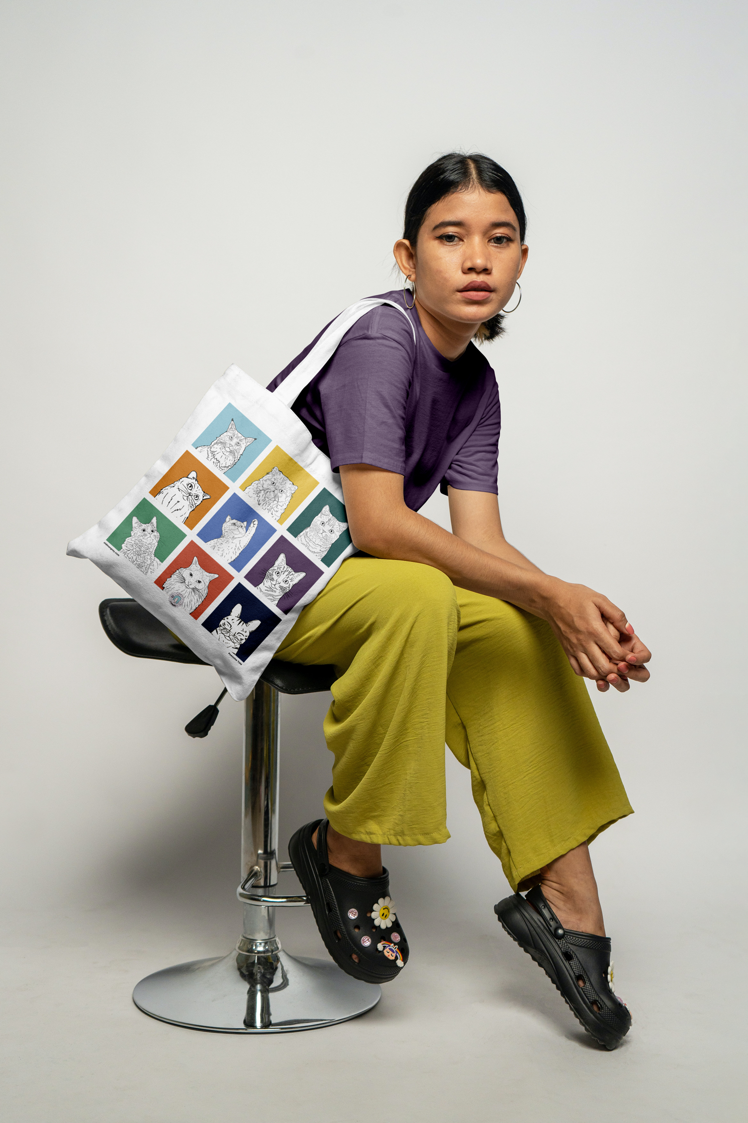 A female model sat sideways on a stool with a tote bag on her shoulder. It has 9 brightly coloured boxes,  with a black & white digital illustration of the head & shoulders of different cat breeds.