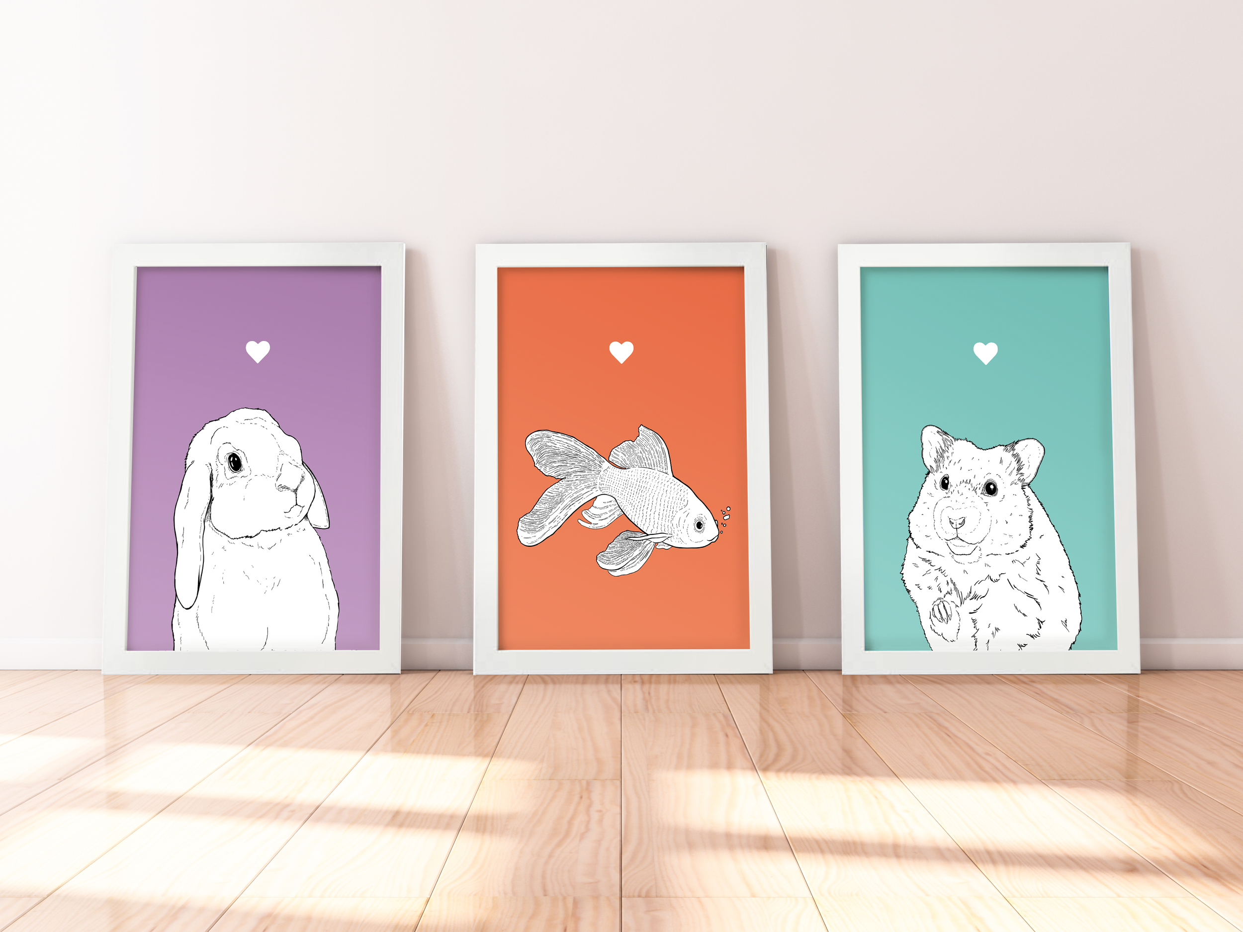 A trio of A4 prints in white wooden frames leaning against a white wall. Each has a black & white digital illustration of a wee pet: Lop Bunny, Goldfish & Hamster