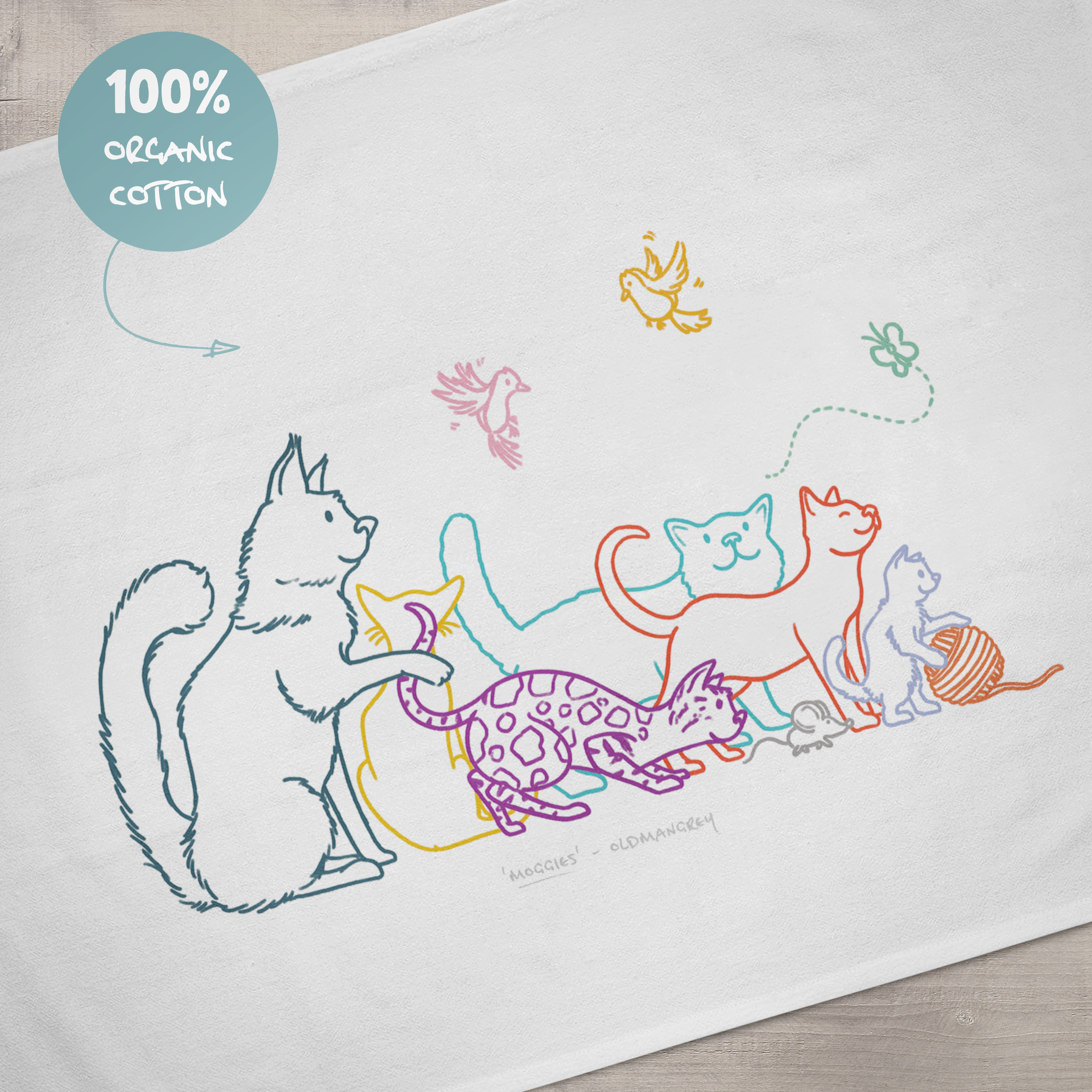 A close up of the design on a large tea towel. It has a colourful digital illustration of 6 cats, 2 birds, a butterfly & mouse. A sign says '100% organic cotton'.