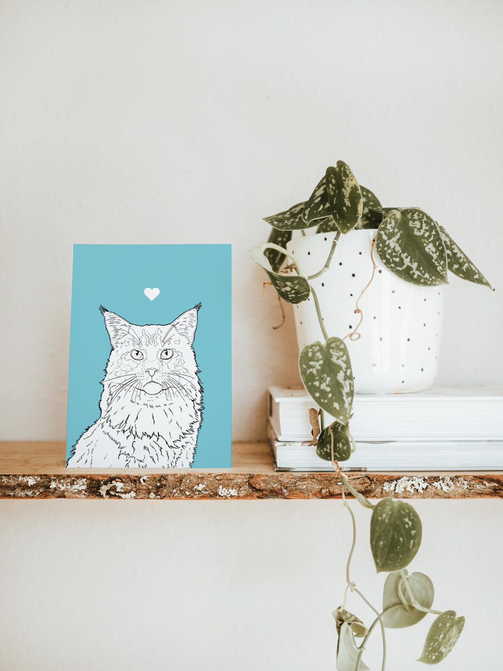 An A6 greetings card & white plant pot on a shelf. The card has a pale blue coloured background, with a black & white digital illustration of the head & shoulders of a Maine Coon cat.