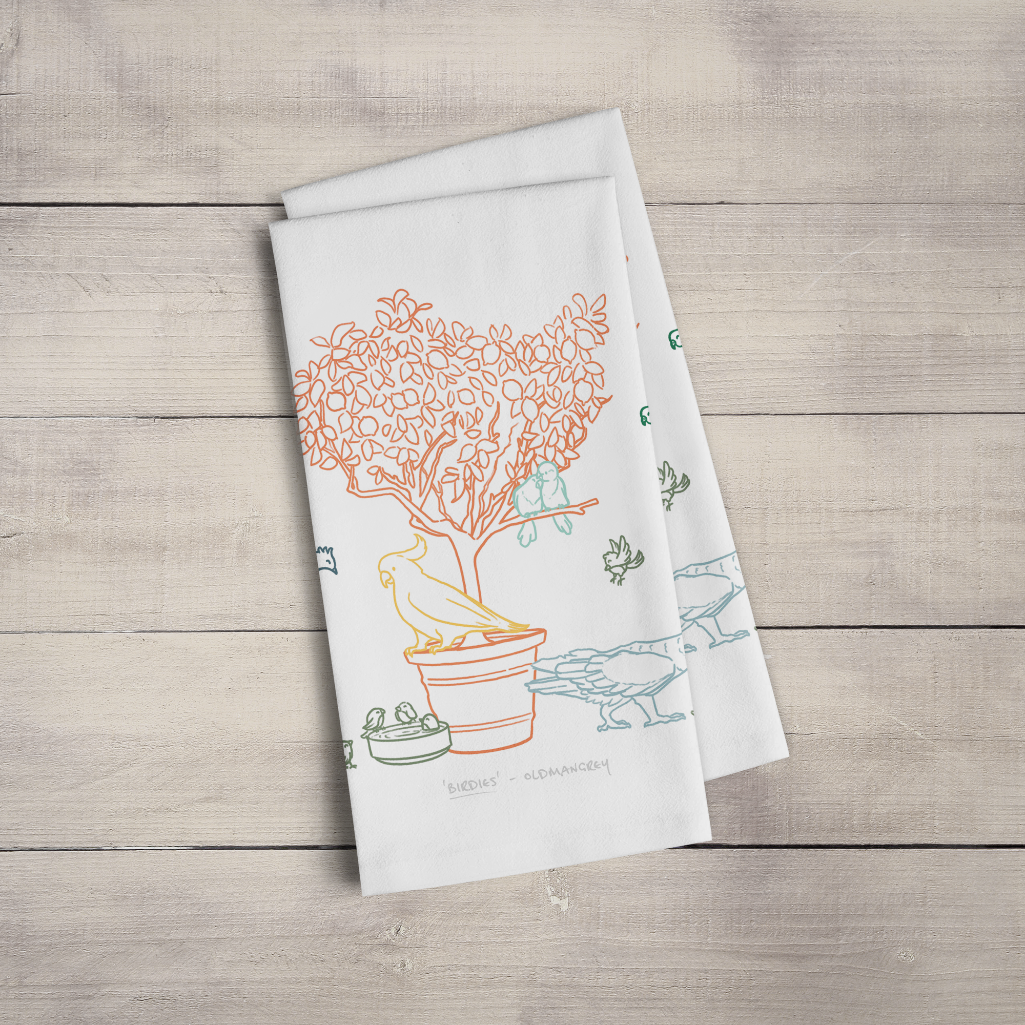 Two large tea towels folded to a rectangle & fanned out, on a wooden surface. Part of the design can be seen - colourful digitally illustrated birds on & around a small tree.