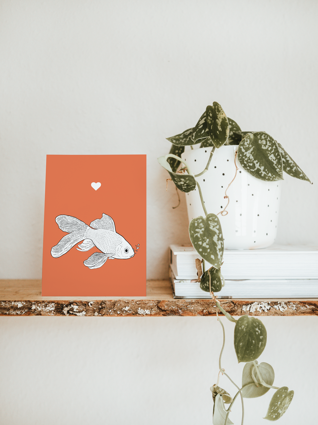 An A6 greetings card & white plant pot on a shelf. The card has a red coloured background, with a black & white digital illustration of a Goldfish.