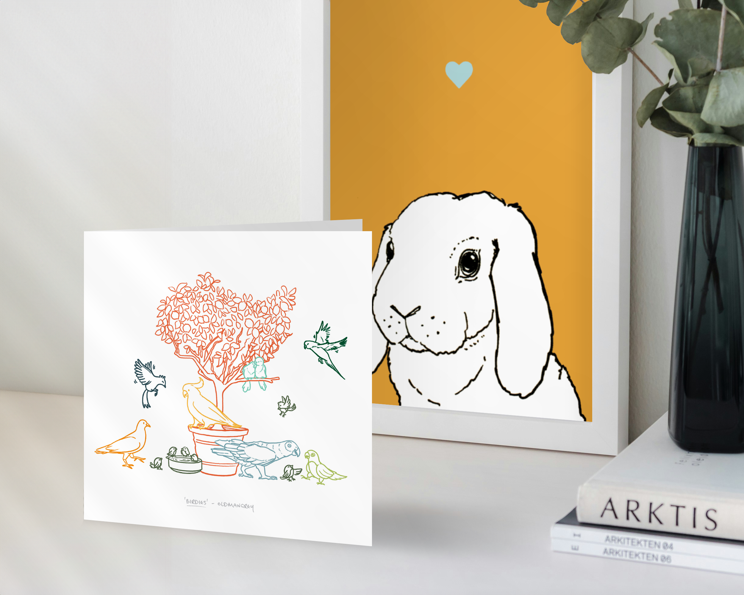 A square greetings card standing in front of a framed rabbit print. The card has a colourful digital illustration of birds on & around a small tree.